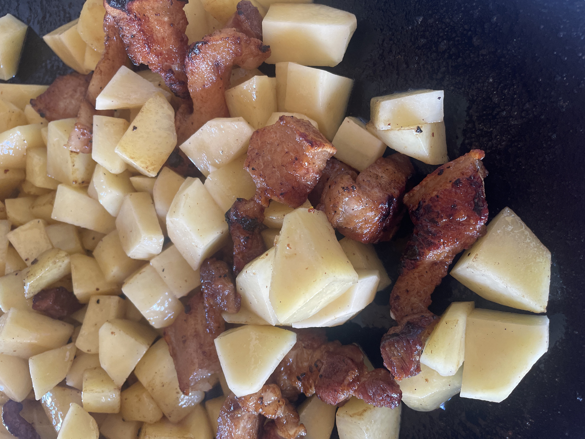 The scene, located in Petrivka, Ukraine, presents a high-angle, close-up view of cooked food. The image primarily features numerous irregularly diced pieces of potato and seared meat. The potatoes are predominantly light yellow to off-white, cut into various polygonal shapes, exhibiting some minor surface browning and a slight oily sheen. Interspersed among the potatoes are chunks of dark reddish-brown meat, characterized by visibly seared surfaces, darker charred edges, and a glistening appearance indicative of rendered fat. These meat pieces vary in size and shape, from elongated strips to more compact cubes. The food rests on a dark, non-reflective surface, likely a cooking pan or skillet, with some isolated dark spots visible on the surface. No individuals or further environmental details are discernible.
