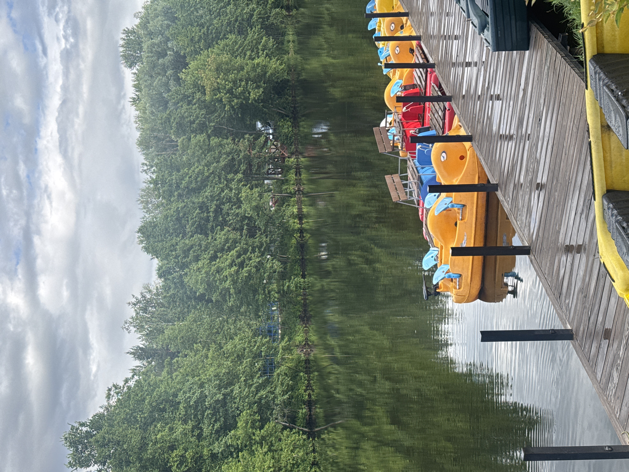 The image depicts a waterfront scene at an unspecified location in Sarov, Russia. The foreground features a wooden dock with several colorful pedal boats moored alongside it. These boats are predominantly yellow and red, with blue accents. The dock appears wet, suggesting recent rain. In the mid-ground, a calm body of water reflects the lush green trees that line the opposite bank. The background is dominated by a dense forest, with the sky above showing a mix of clouds and patches of blue. No people are visible in the scene.