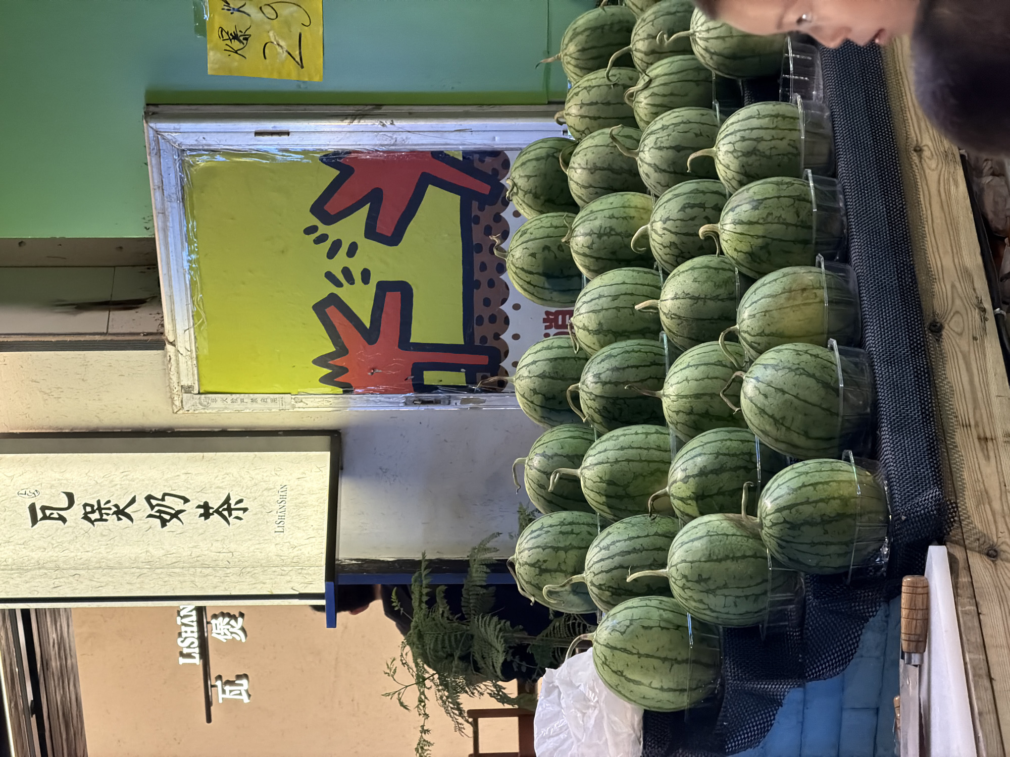 The scene depicts a collection of small, round, striped watermelons neatly arranged in clear plastic cradles on a black mesh surface. A person, partially visible in the upper right corner of the frame, appears to be examining the watermelons.
In the background, there's a colorful yellow sign with a red and black cartoon figure, possibly an advertisement. To the left of the watermelons, a vertical sign with Chinese characters is displayed, indicating "Lishan Shan Tea." Another sign below it in English and Chinese also refers to "Lishan". A fern plant is visible behind the bottom-most row of watermelons.
Based on the signage and available information, the location is likely Chongqing High-tech Zone in China. The presence of both English and Chinese text indicates a commercial or tourist-oriented area. The focus of the scene is on the sale of watermelons, and the presence of the individual suggests potential interaction or purchase.