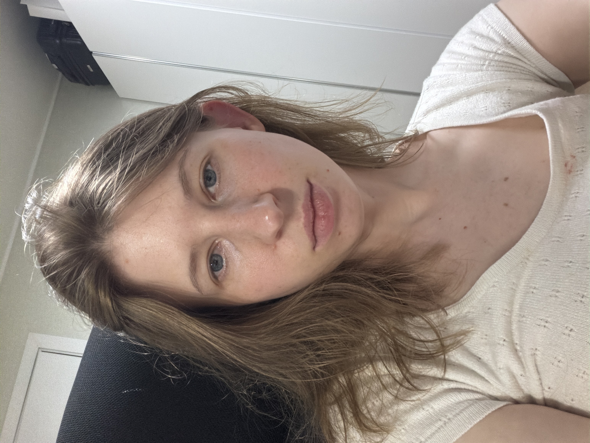 A woman with light brown hair and blue eyes is pictured in a close-up, head-and-shoulders shot. She is wearing a light-colored, short-sleeved, knitted shirt. Her skin appears fair with some redness on her cheeks and a few small brown spots. Her hair is parted in the middle and falls loosely around her face. She is looking directly at the camera.
The background is predominantly white, with a dark object, possibly a suitcase, visible on top of a white cabinet or shelf in the upper right corner of the image. To the left, a dark, textured surface, likely the back of a chair, is partially visible. The setting appears to be an indoor environment, possibly an office or a room with furniture.
The image was taken in Dongducheon-si, South Korea.