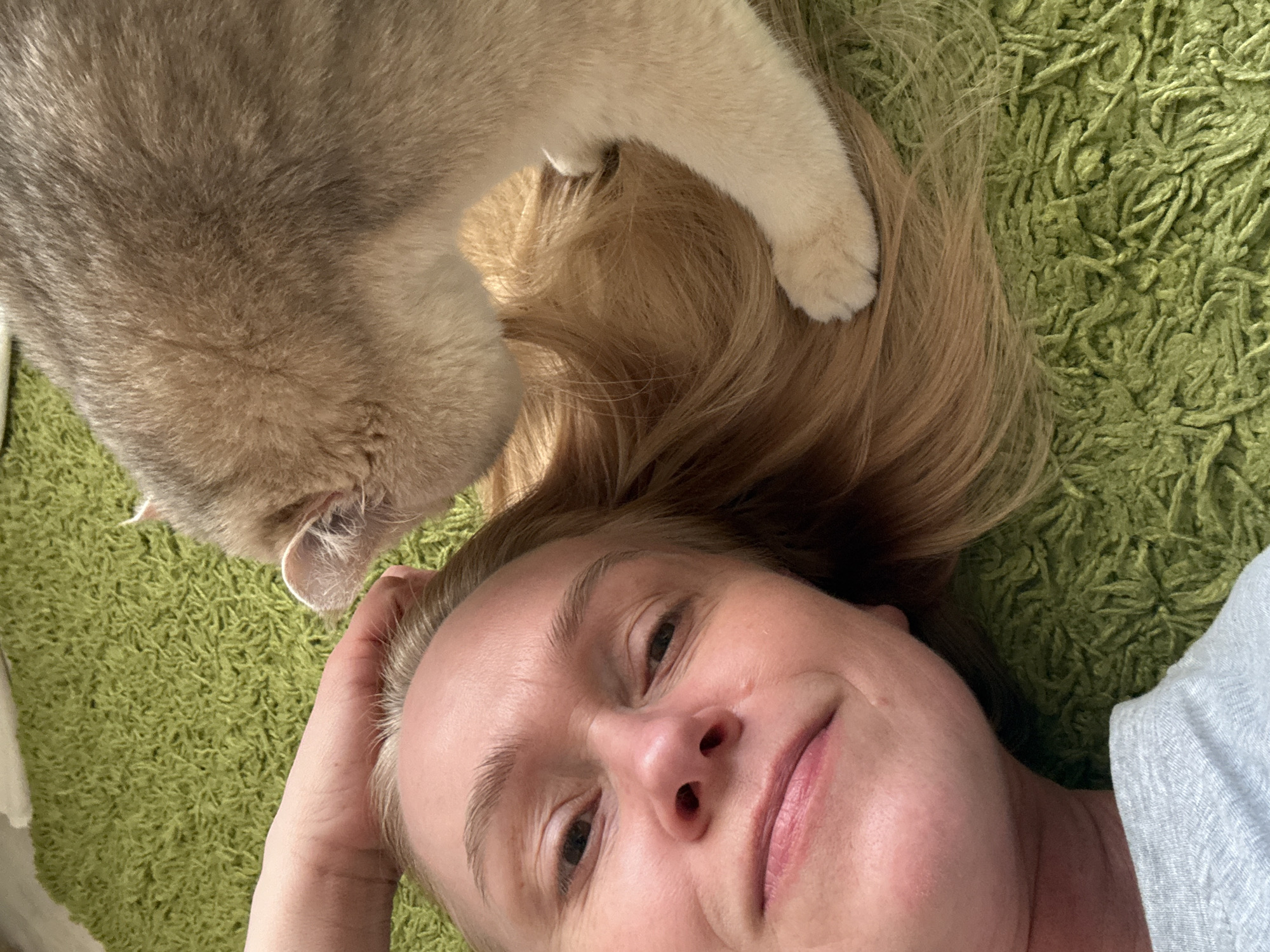A selfie-style shot shows a woman lying on a green shag rug. She is looking up at the camera with a slight smile. A tabby cat is perched above her, with its paw resting on her blonde hair. The background consists solely of the green rug. The location is possibly in Chaikovsky, Russia, but there are no features visible that provide explicit confirmation.