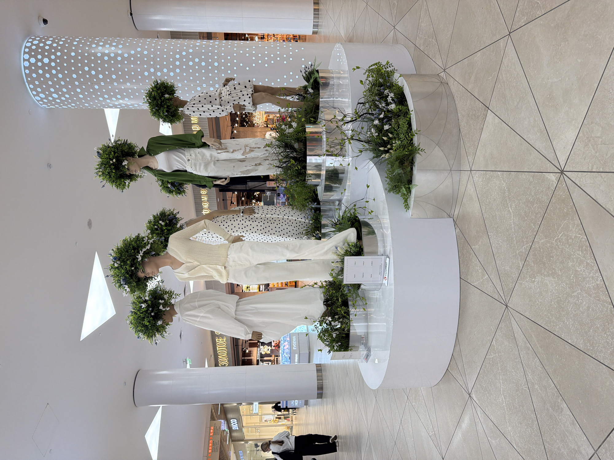 The image shows a shopping mall in Kazan, Russia, with a multi-tiered display of mannequins. There are four female mannequins on the display. The mannequins are each wearing a wreath of flowers on their heads. The mannequins are dressed in modern clothing. The display is built on a circular platform and is surrounded by glass panels and planted greenery. The interior architecture of the mall features geometric tile patterns on the floor and cylindrical support pillars. Shoppers are seen in the background, walking around the mall. Storefronts with visible signage, including shops named "ZOLOTOE" and "EVGENIA" are visible.