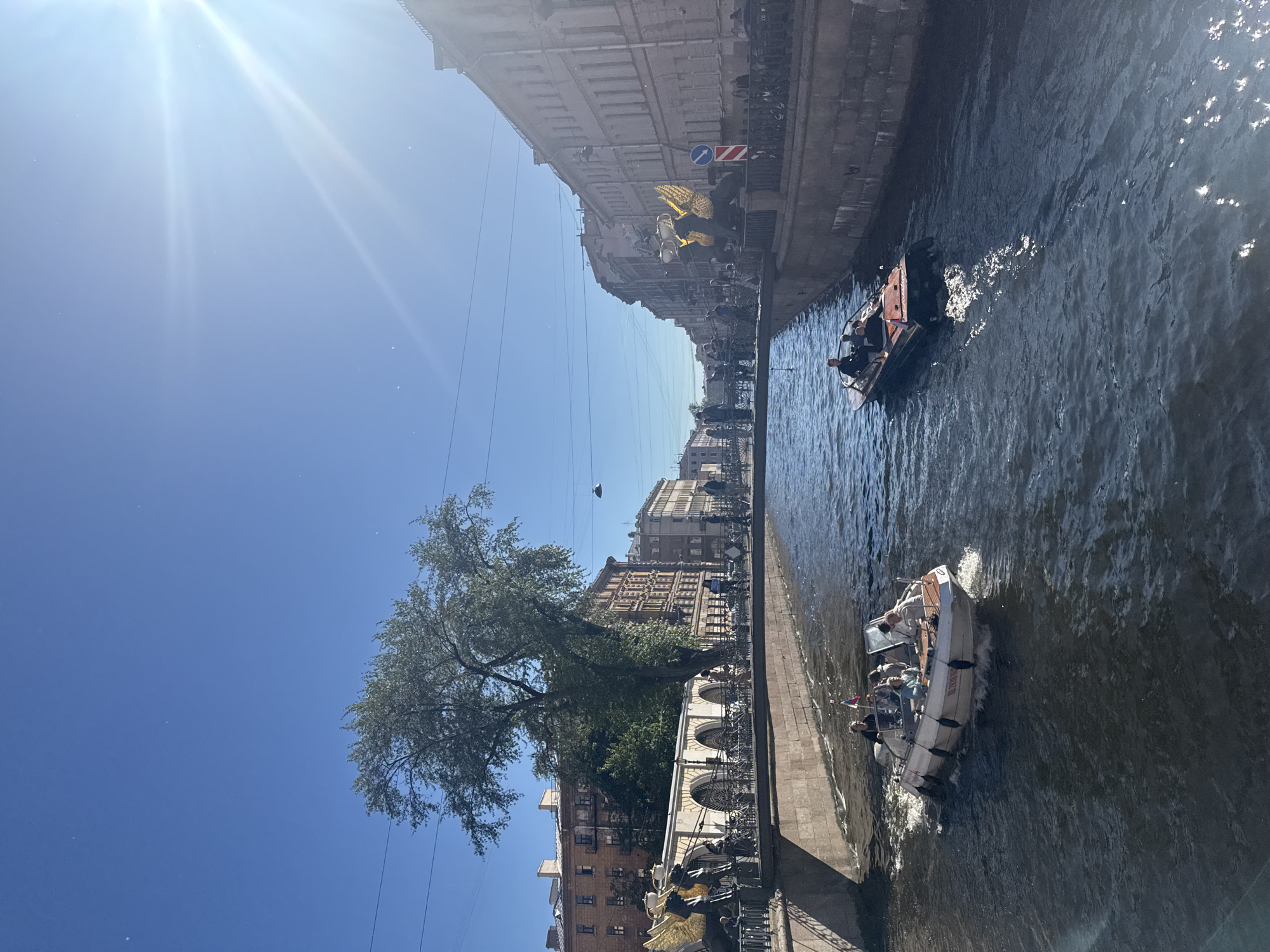 This is a vertical, high-angle shot of a canal in Saint Petersburg, Russia. The sun is bright and high in the sky, suggesting it is midday and the weather is clear. The canal is lined with ornate buildings that appear to be historical architecture. A large tree with green leaves is visible on the left bank, casting some shade.
There are two boats on the canal. In the foreground, a small, gray inflatable boat with an outboard motor is carrying several passengers. Some of the passengers appear to be tourists, and they are all looking in different directions. The boat is moving at a moderate speed, creating a wake. In the background, another similar boat is also moving.
The overall mood of the image is relaxed and pleasant, suggesting a leisurely day of sightseeing. The presence of boats and tourists indicates this is a popular tourist area. The architecture of the buildings, with their detailed facades and arched windows, hints at the city's rich history and cultural significance. The canal itself appears to be a navigable waterway within the city, likely a popular route for boat tours.