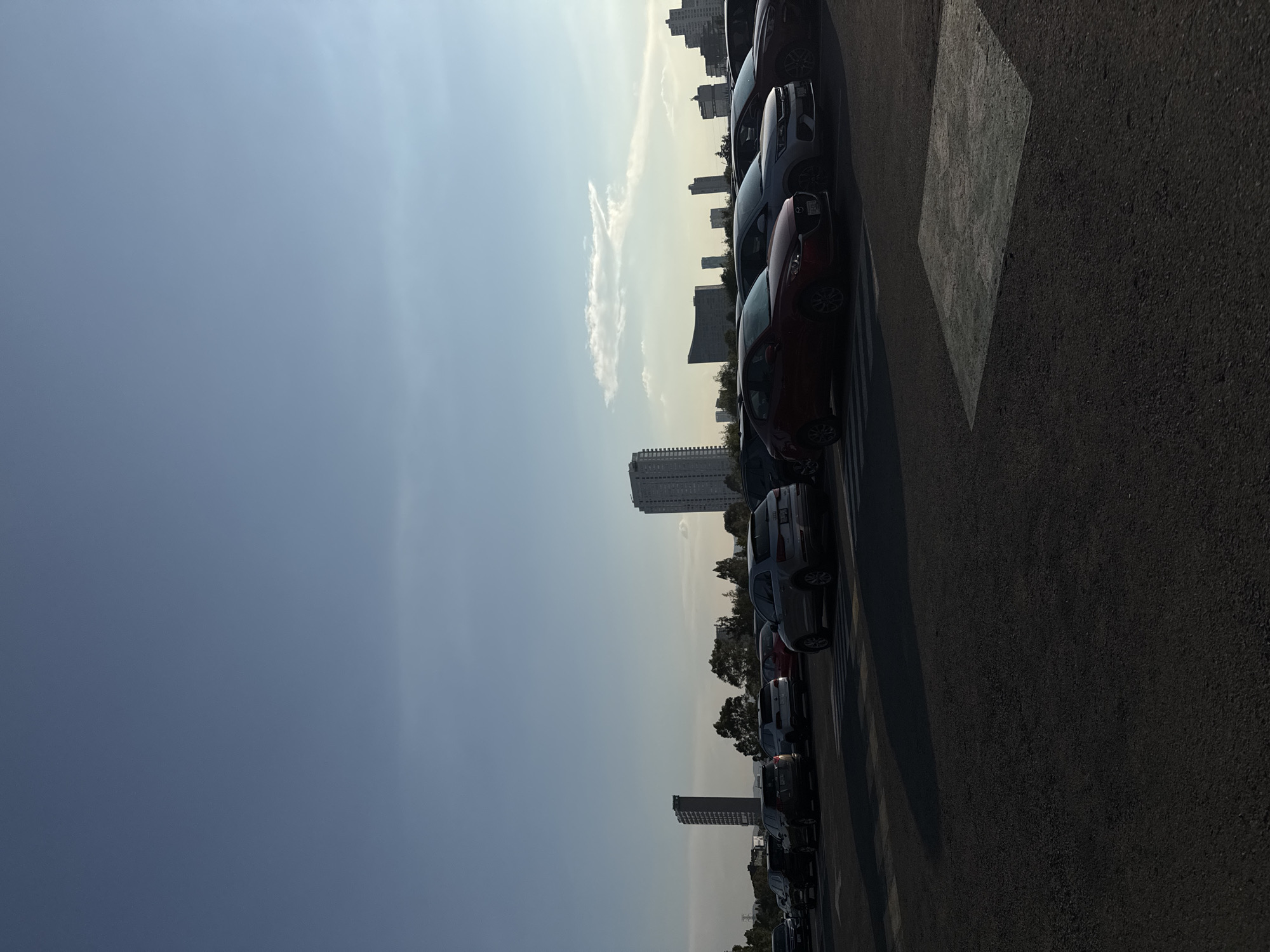 The image shows a row of parked cars along a street in Puebla, Mexico. The cars are parked along a parking lot next to a street. In the background, several buildings of varying heights can be seen, suggesting an urban environment. The sky is clear and bright, with light clouds near the horizon. The photo appears to have been taken during the day with strong sunlight, as indicated by the shadows. The surface is asphalt.