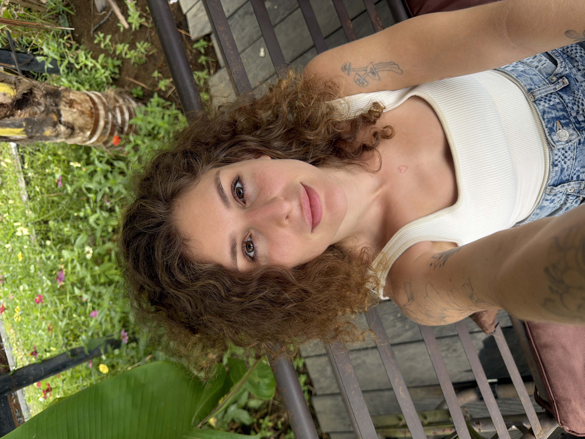 An overhead selfie of a young woman with curly brown hair is captured from a high angle. She is seated on a wooden bench with dark metal slats and a brown cushioned seat. She wears a white ribbed tank top and distressed denim jeans. Visible tattoos adorn her right arm, one appearing to be mushrooms, and a small red heart tattoo is on her upper chest. She is outdoors, with lush green foliage and a variety of colorful flowers in the background. To the left of the frame, a large, vibrant green leaf is prominent. Behind her, a portion of a palm tree trunk with yellow and black markings is visible. The ground appears to be dark soil with some paving stones. The lighting suggests it is daytime. The location is identified as Mengwi, Indonesia.