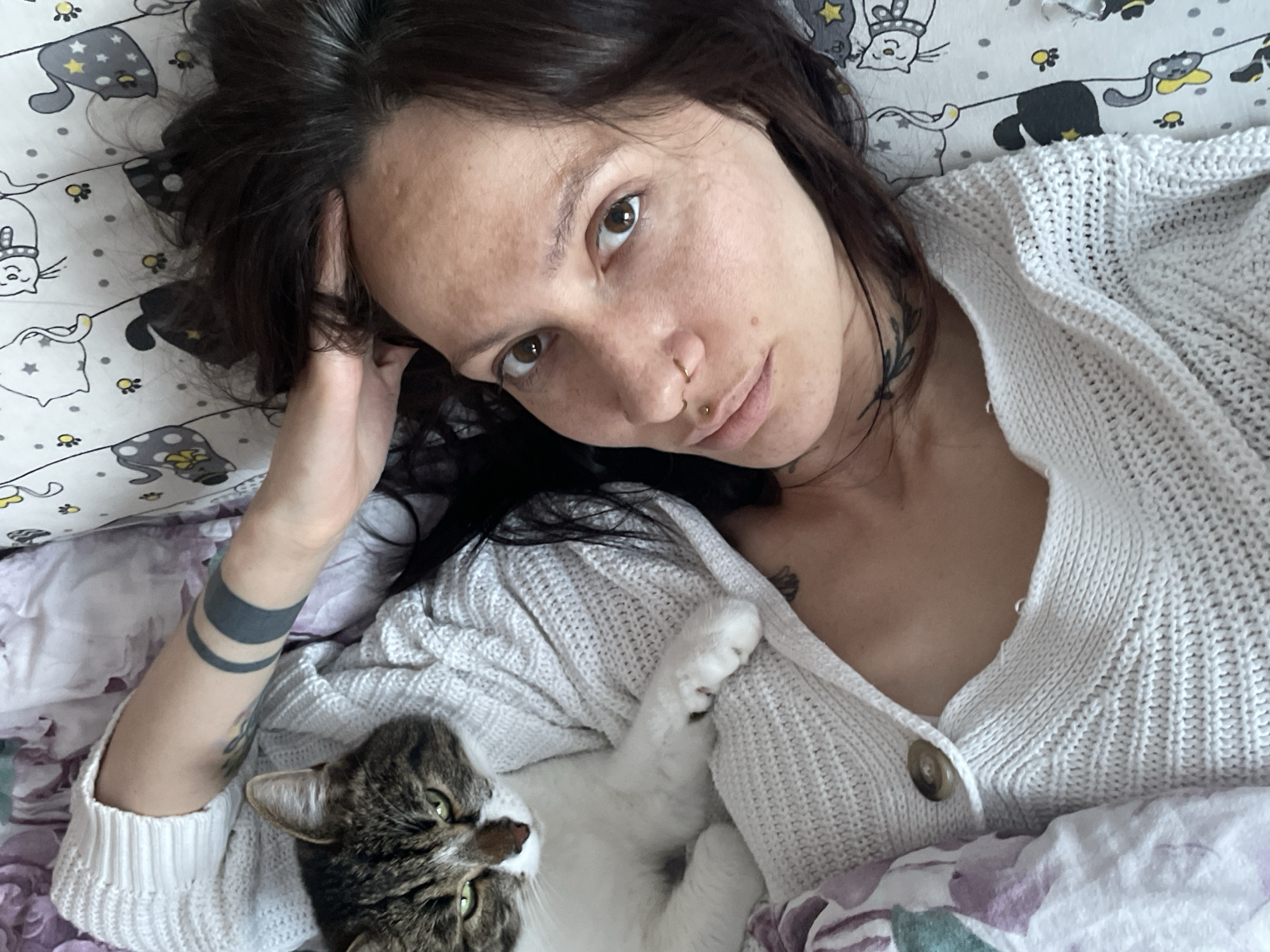 A woman with dark hair and light skin lies in bed, looking at the camera. She has a piercing in her nose and lip, and tattoos are visible on her neck, chest, and left arm. She is wearing a white, knitted cardigan. A cat with green eyes and brown and white fur rests on her chest. The bedsheets have a pattern of cats and moons. The image was taken indoors. The location in the city of Izhevsk, Russia cannot be confirmed from the provided image.