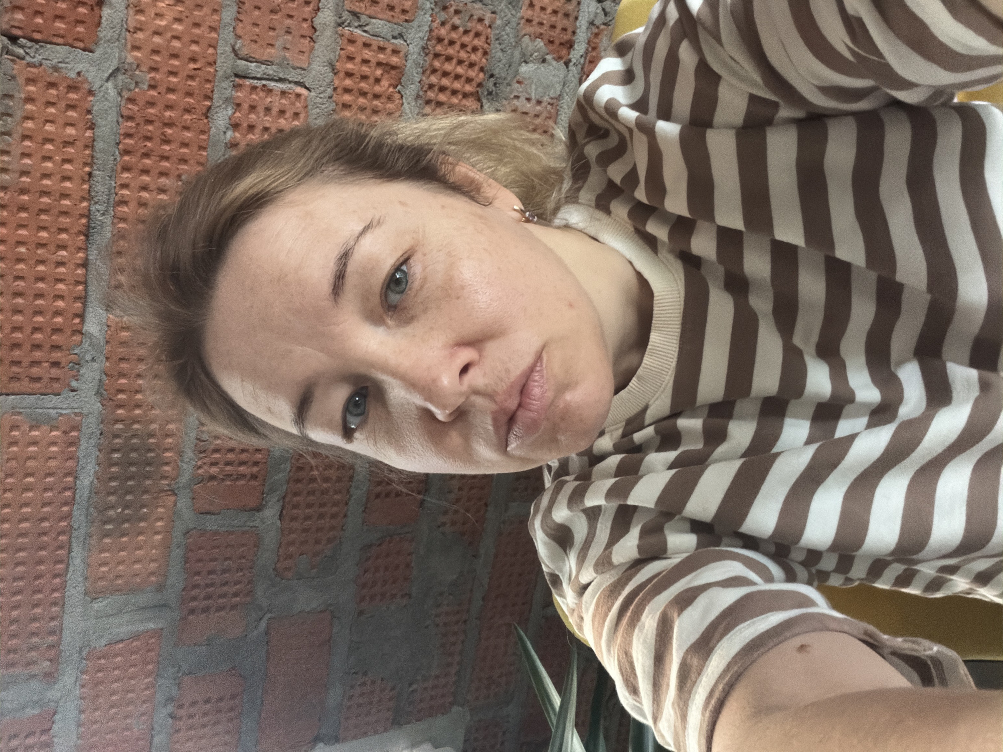 A woman is posing for a selfie indoors, with a red brick wall in the background. She is wearing a striped brown and white shirt. The location is possibly in Pervomaysky, Russia.