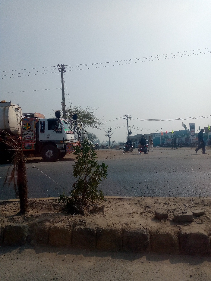 The image depicts a daytime street scene in Dijkot, Pakistan, featuring a mix of vehicles, pedestrians, and local infrastructure under a clear sky.
In the foreground, a low dirt median or curb separates the viewer from the main road. Two small trees or bushes grow from this median, along with the visible trunk of a palm tree on the left. The road itself is paved and appears busy.
A prominent subject is a large white tanker truck on the left side of the road. It is heavily decorated with colorful, traditional Pakistani "truck art," including intricate patterns and some illegible Urdu script or calligraphy. The truck appears to be either stationary or moving slowly. Further down the road, several motorcycles are visible, with riders on them. A couple of individuals are also seen walking across or alongside the road in the mid-ground, one particularly distinct on the right side.
The background features a building with text on its facade, clearly visible as "IQRA FEDERAL COLLEGE FOR GIRLS & BOYS." This building has multiple flags or banners displayed in front of it. Tall utility poles with numerous overhead wires crisscross the frame, indicating electrical or communication infrastructure. The overall weather appears sunny and clear, casting distinct shadows. The scene captures a typical bustling street environment in the region.