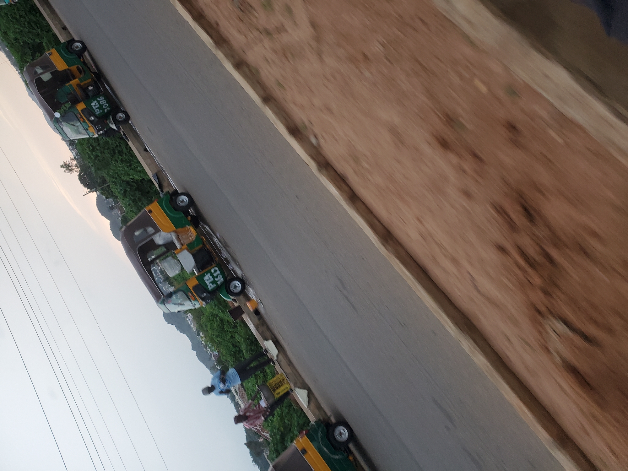 This is a low-angle, motion-blurred photograph taken in Bukuru, Nigeria. The image depicts a street scene with three auto-rickshaws, commonly known as Keke Napep, on a paved road. The Keke Napep vehicles are primarily green and yellow. The first Keke Napep, on the far left, is partially visible and is facing away from the camera. The second Keke Napep is in the center of the frame, with "PTA 7086" visible on its side. It is also facing away from the camera and appears to be stationary. Two men are standing on the sidewalk next to this Keke Napep. One man, wearing a blue t-shirt and dark pants, is standing and looking at his phone. The other man, wearing a red shirt, is near a yellow crate and appears to be interacting with the driver or passenger of the Keke Napep. The third Keke Napep is on the right side of the image, with "PTA 1613" visible on its side. It is also facing away from the camera and is in motion. The background consists of lush green vegetation and a hazy, hilly landscape, typical of Bukuru. The sky is overcast. The foreground is dominated by a dirt shoulder and the edge of the paved road.