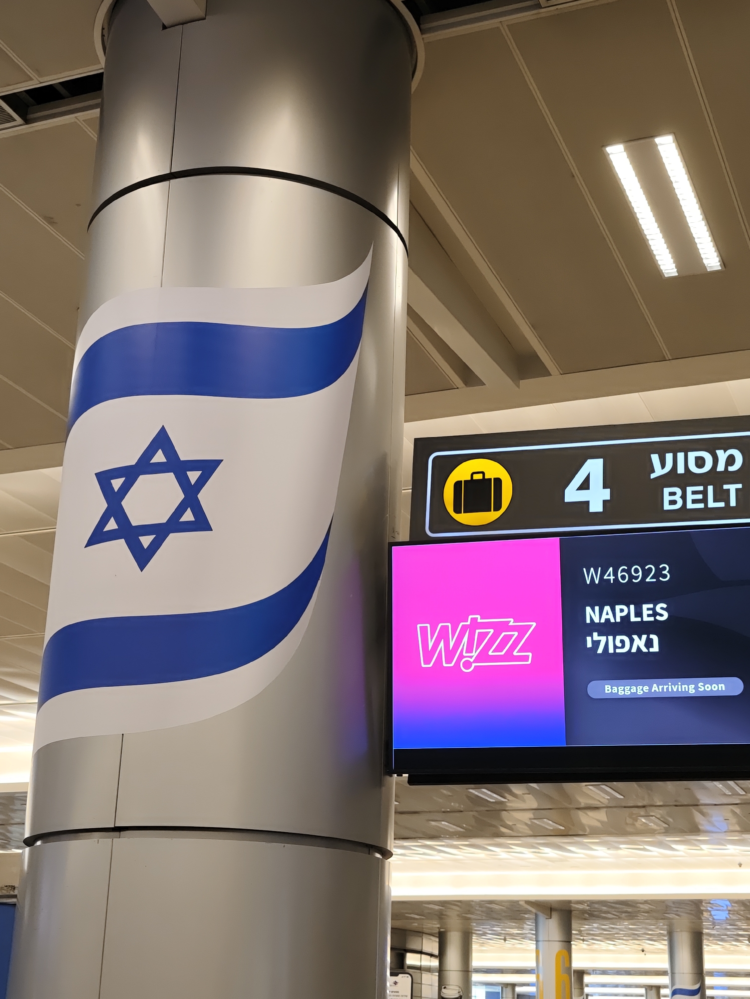 An airport interior scene features a large, cylindrical pillar decorated with a prominent Israeli flag design. To the right of the pillar, a digital display screen shows flight information. The screen is divided into two sections. The top section, darker in color, displays the number "4" in large white digits, accompanied by a yellow briefcase icon and the Hebrew word for "Baggage Claim" above the English word "BELT". Below this, a pink and purple section displays the "Wizz" airline logo. Further down, flight information includes the flight number "W46923", the destination "NAPLES" in English, and "נאפולי" in Hebrew. The very bottom of the screen shows the text "Baggage Arriving Soon". The environment appears to be a modern airport terminal with suspended ceilings and recessed lighting fixtures.