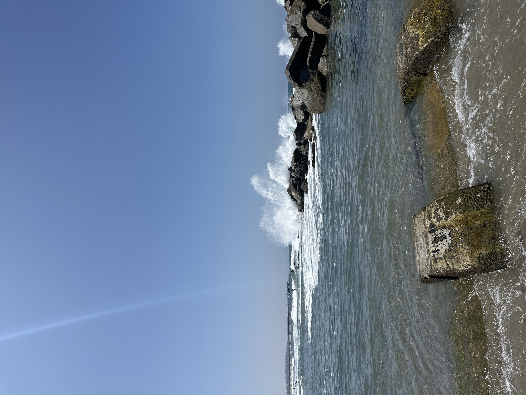 This image shows a coastal scene in Los Angeles, California. The foreground features a sandy beach with two rectangular concrete blocks partially submerged in the water, with waves lapping around them. The water is a clear blue-green, with some white foam from the waves. In the middle ground, a rock jetty juts out into the ocean. Waves are crashing against the rocks, creating white spray. Several surfers can be seen in the water, navigating the waves. In the background, a long pier extends into the ocean, with a few people visible on it. The sky is a clear, bright blue. The overall impression is of a sunny day at the beach with active ocean conditions.