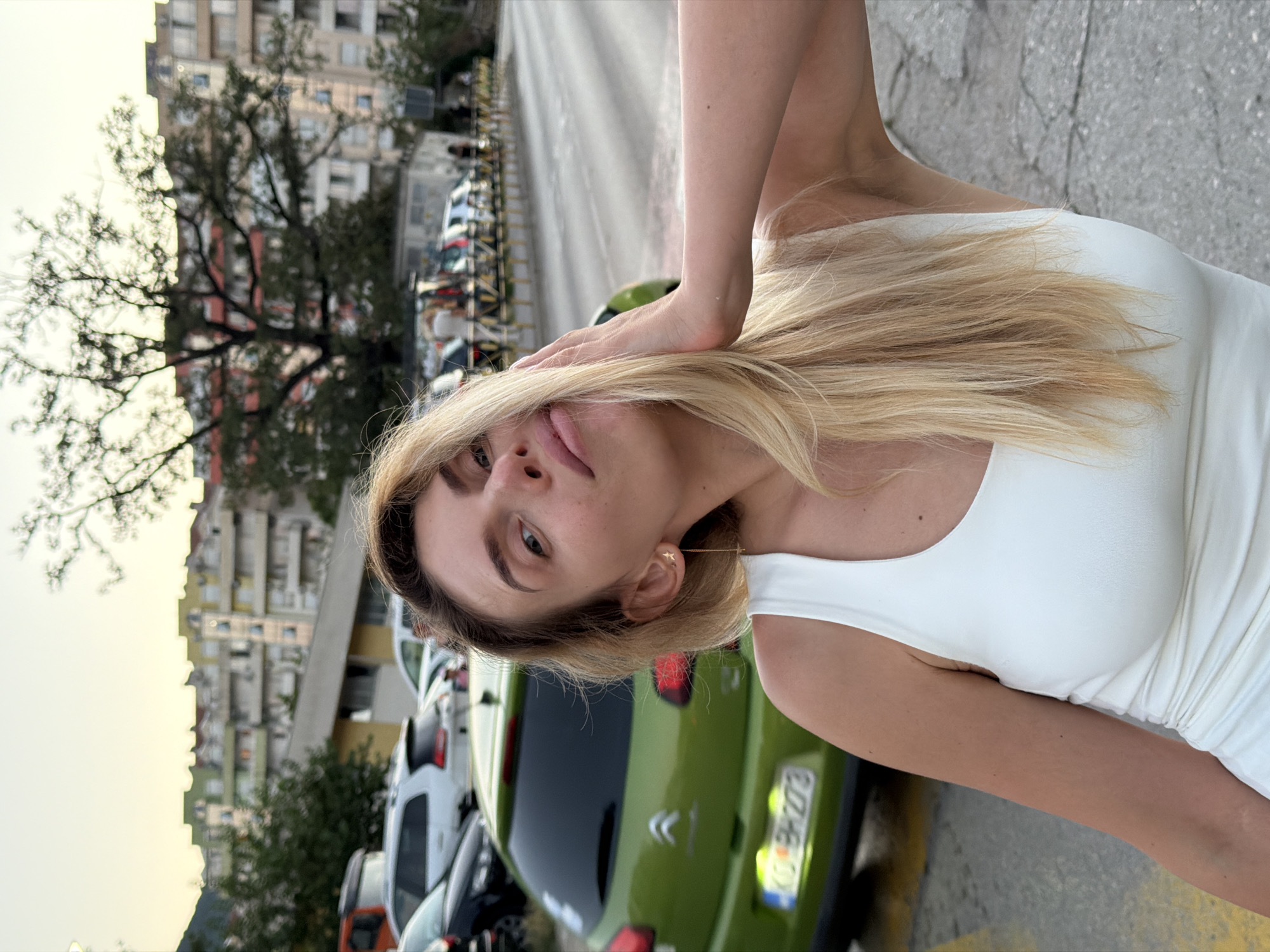 A woman with blonde hair and a white tank top is in the foreground, with her left hand touching her hair and her face turned towards the camera. Behind her, a green car is parked on a street, and in the background, there are buildings and trees. The scene appears to be in Budva, Montenegro, possibly near the Old Town, given the architecture of the buildings.