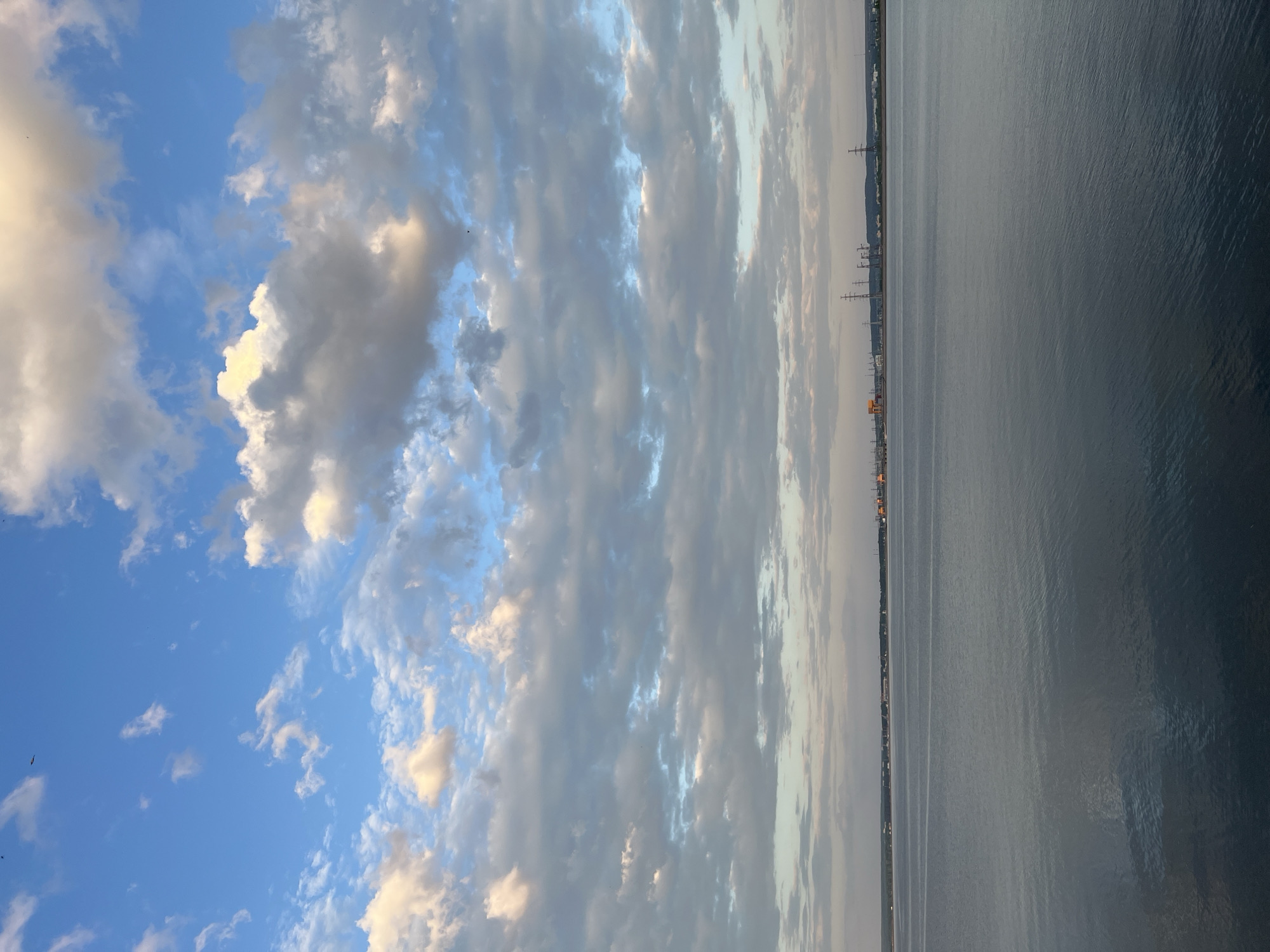 The image depicts a horizontal view of a scene with sky, clouds, and water. The left portion of the image features a blue sky with cumulus clouds that range in color from bright white to grey. The right portion of the image shows a body of water that appears grey and calm. On the distant shoreline, a line of buildings and structures is visible, along with utility poles and power lines. The location is identified as Noviy, Russia. No people are visible in the image. The interaction is between the elements of nature and the built environment along the shoreline.