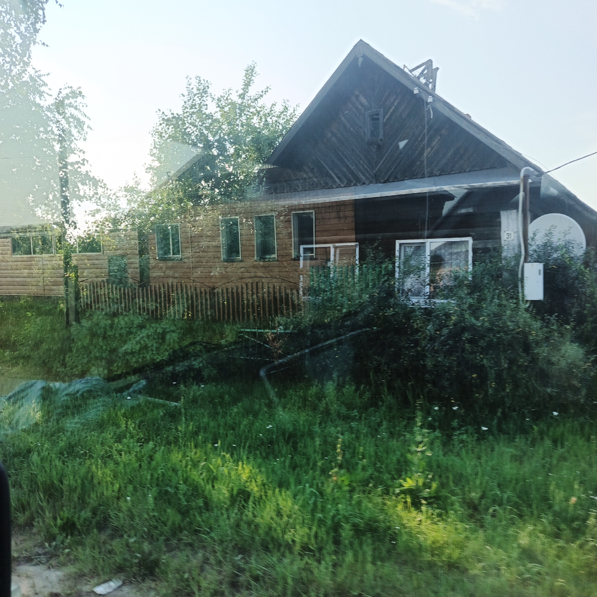 The image shows a rural scene, likely a residential property, under a bright, possibly hazy sky. The primary focus is a wooden house, constructed with horizontal wooden planks for the main body and vertical planks for the gabled roof portion. The house features several windows, some with white frames. To the right of the house, there is a satellite dish and an electrical box mounted on the exterior wall. A wooden fence with vertical pickets runs in front of the house, partially obscured by lush green vegetation and tall grass in the foreground. Trees and foliage are visible behind and to the sides of the house. The scene appears to be taken from a moving vehicle, as indicated by a slight blur and reflection on the left side of the image. The environment is consistent with a rural setting in Kambarka, Russia, characterized by traditional wooden architecture and abundant greenery. There are no people or discernible actions taking place in the image.