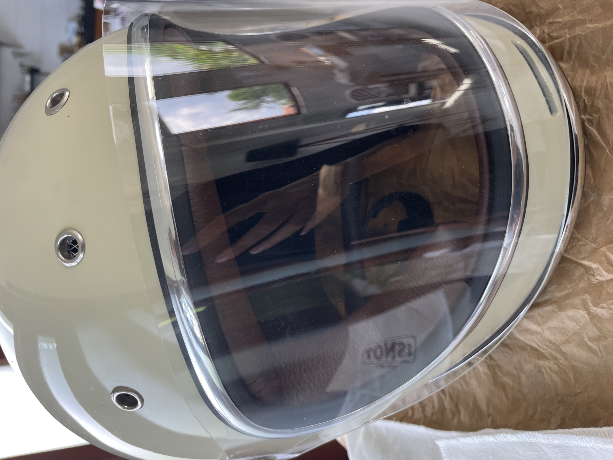 A motorcycle helmet with a clear visor sits on crumpled brown paper, possibly packaging material. The helmet is light cream in color and has metal vents on the side. The visor reflects the outside environment, showing hints of trees and buildings. A hand is visible in the reflection. The background suggests an indoor setting near a window. There is no indication of the location.