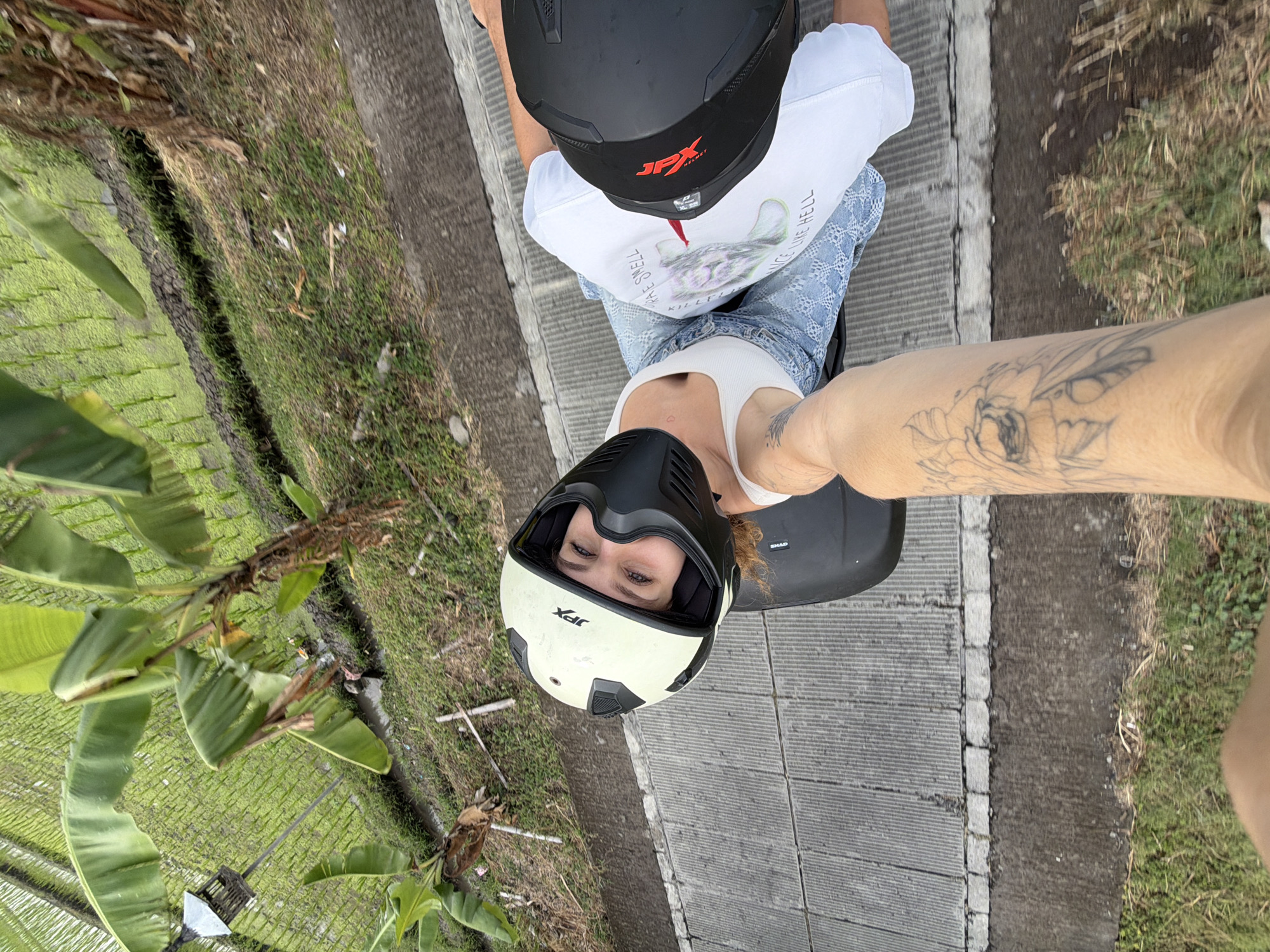 A high-angle, POV shot captures a couple riding a scooter through a rural area with rice paddies in North Kuta, Indonesia. The woman, in the foreground, is wearing a white tank top, denim shorts, and a white and black helmet, with a black face mask. Her eyes are open, and she is looking at the camera. Her left arm, with a tattoo of a flower, is extended towards the camera. The man, in the background, is wearing a white t-shirt with a graphic design and a black helmet. They are traveling on a paved road next to a lush green rice paddy. The sunlight is bright, casting shadows.