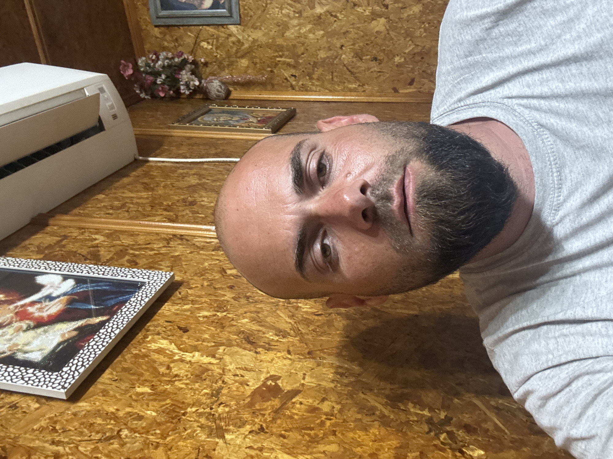 This image captures an indoor scene, set in the city of Sarnevo, Bulgaria. The central subject is an adult male, bald, with a dark brown, full beard covering his chin and jawline. He has dark eyebrows and brown eyes, looking directly forward with a neutral expression. His skin shows natural texture and some redness on the cheeks and forehead. He is wearing a light grey, heathered, short-sleeved t-shirt with a visible ribbed crew neck.
The background consists of walls uniformly covered with Oriented Strand Board (OSB) panels, displaying a distinct, textured, warm yellowish-brown surface. In the upper left quadrant, a framed picture is mounted on the wall. Its frame is white with a black stippled pattern, enclosing what appears to be a classical or religious painting depicting multiple figures in light and dark garments.
In the upper right quadrant, a white indoor split-system air conditioning unit is mounted high on the wall, partially open with its dark vent visible. Below the air conditioner, a small, artificial floral arrangement containing pink and white flowers is attached to the wall. Further down and to the right, a small, dark-framed religious icon or painting is visible. To its right, another vertically oriented framed picture with a bluish-grey frame is partially visible. The scene is illuminated by artificial light, creating soft shadows.