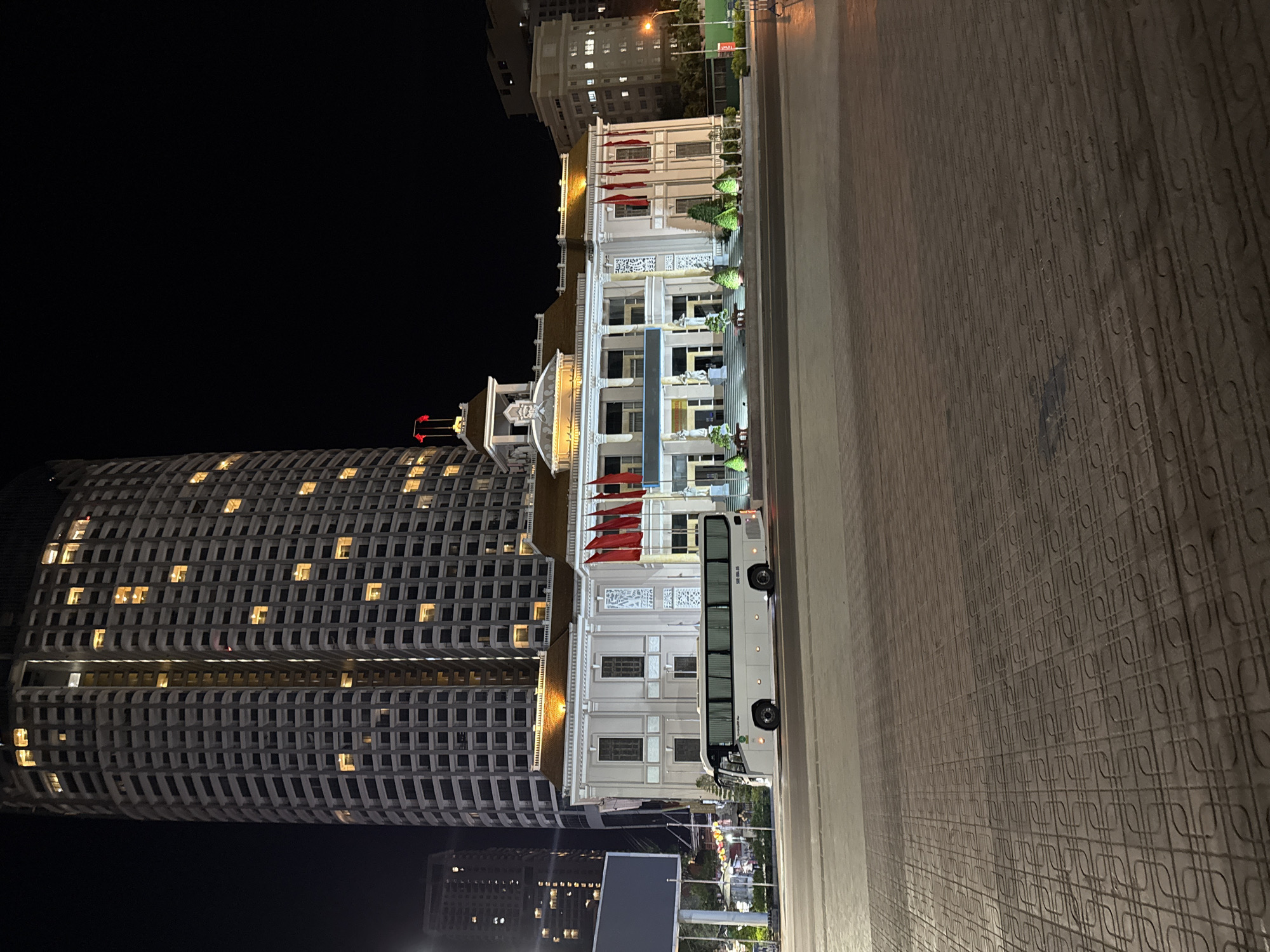 Captured at night, the scene showcases a modern building with a unique grid-like facade and lit windows, towering alongside the illuminated Nha Trang City Hall in Nha Trang Ward, Vietnam. Red flags adorn the City Hall. A bus is parked in front of the building. The pavement in the foreground features a distinct textured pattern. The sky is dark, with other distant buildings barely visible.
