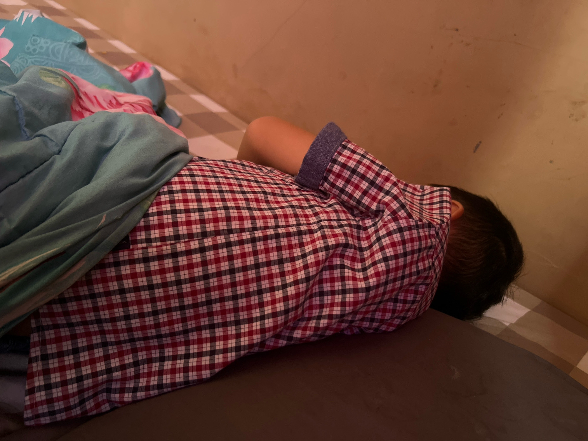 A person is sleeping in a bed, covered by a blue and floral patterned blanket. They are wearing a red and white checkered shirt with a blue cuff. The bed has a checkered bedsheet and is positioned against a wall. The lighting suggests it is nighttime or indoors. The setting appears to be a bedroom. No visible text is present.