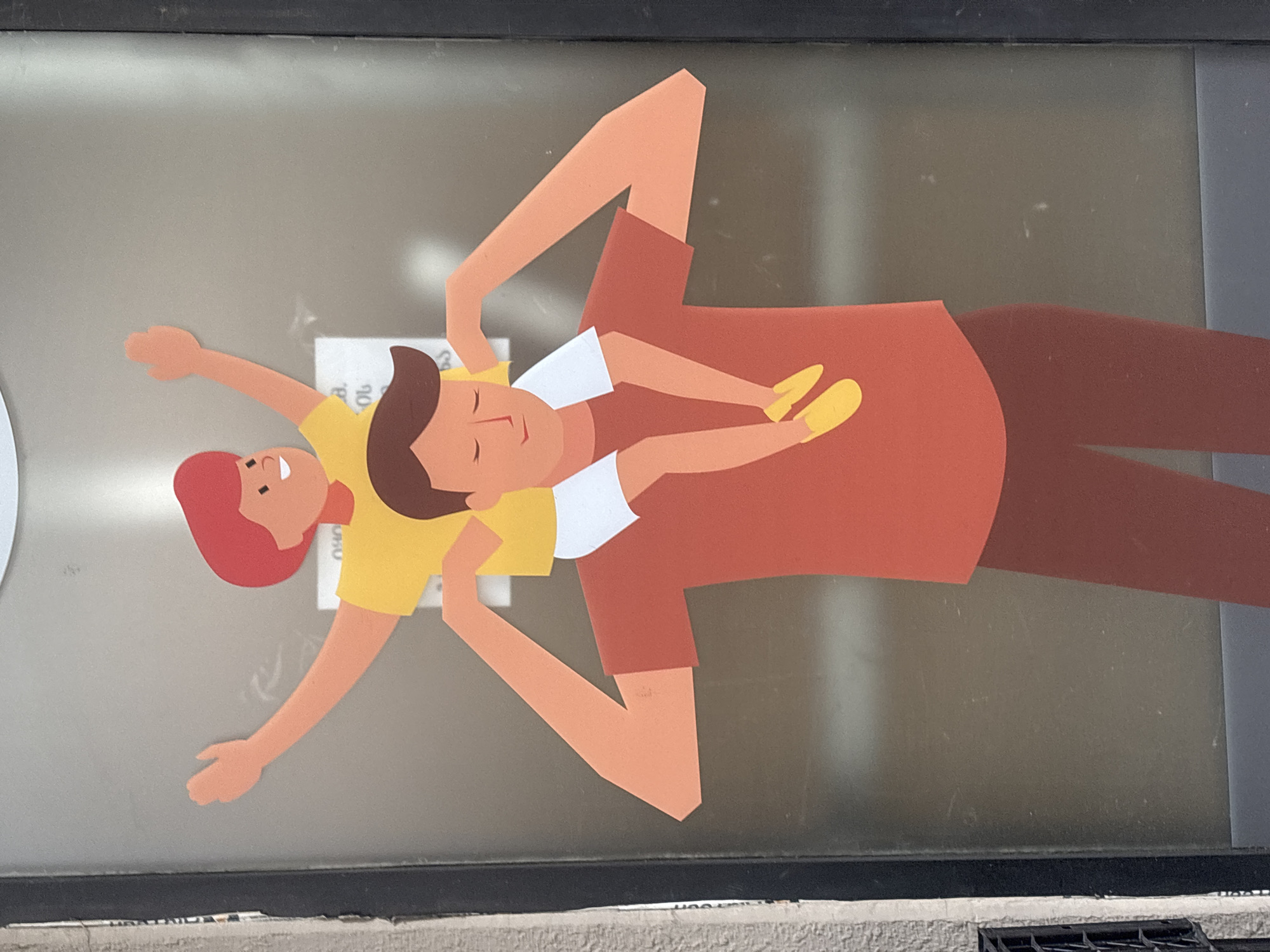 A sticker depicting a father and two children is adhered to a window. The father is wearing a rust-colored shirt and pants, while one child is perched on his shoulders with a white shirt and yellow shoes. The other child is lying next to the father with a yellow shirt and red hair. The sticker appears to be located in Tbilisi, Georgia.