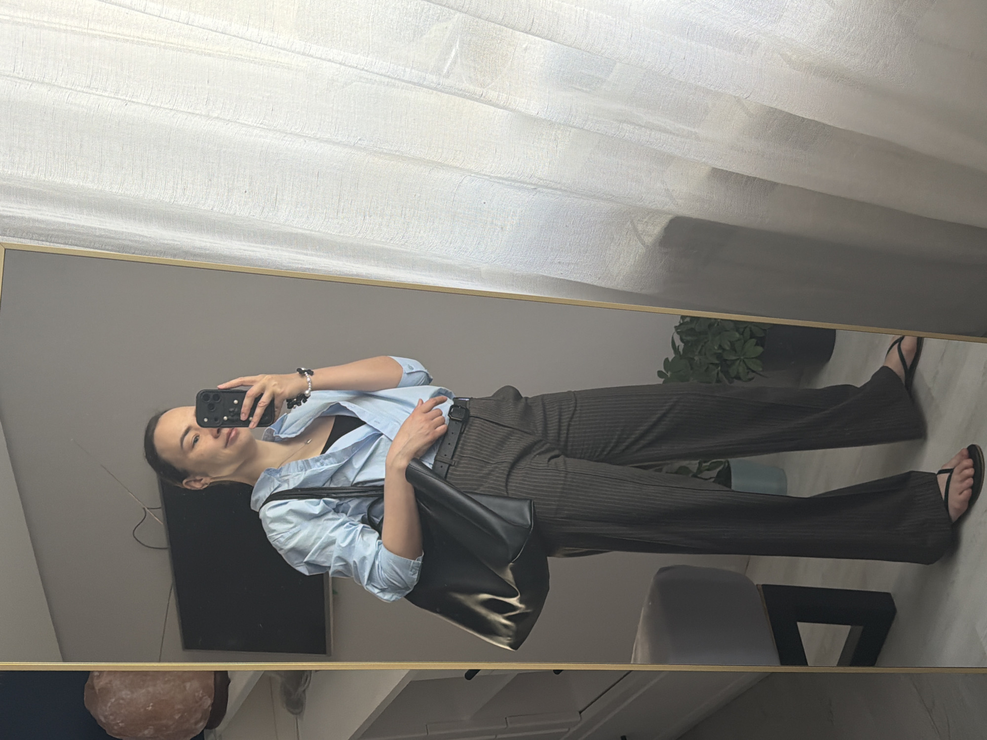 A young woman is taking a selfie in a mirror. She is wearing a light blue button-down shirt, a black top underneath, pinstriped pants, and black sandals. She has a black purse slung over her shoulder and is holding a smartphone in her right hand.
The background features a window with white curtains, a dark-colored television, a Himalayan salt lamp, a potted plant, and a light grey chair with a geometric base. The location is likely an indoor residential setting, though the exact city in Russia (Chaikovsky) is not discernible from the image alone.