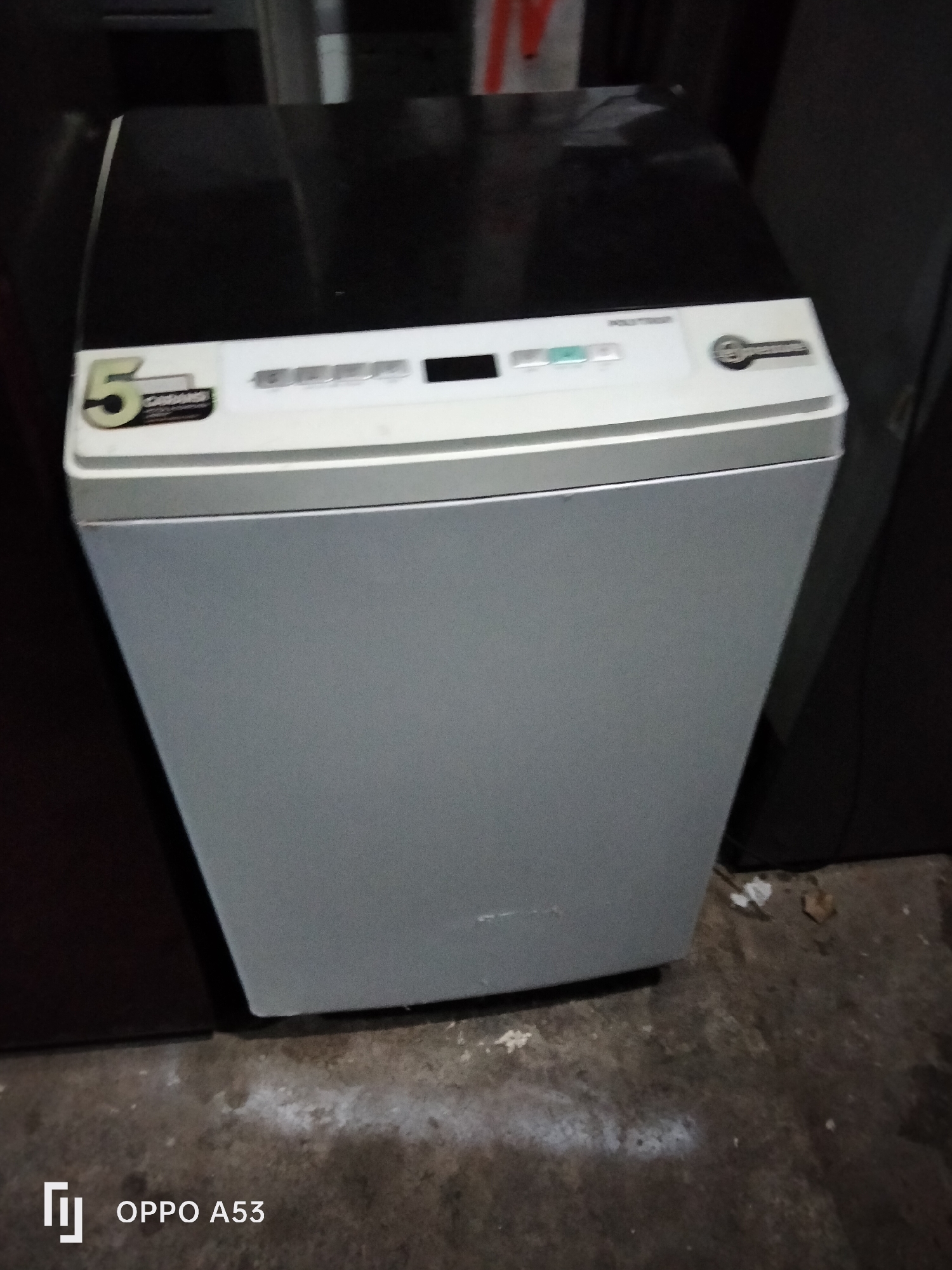 The image shows a top-loading washing machine with a silver body and a black lid. The washing machine is positioned in what appears to be a storage area or a room with a concrete floor. There are hints of other appliances or furniture in the background, with dark vertical lines suggesting the presence of other large items. The floor has some dust and debris, with faint white markings and scattered small items like leaves or paper. On the lid of the washing machine, there are control buttons and a digital display. A sticker with a "5" and the word "GARANSI" (warranty in Indonesian) is visible on the left side of the lid. Another sticker with a logo and the word "POLYTRON" is present on the right side. The bottom right corner of the image shows the text "OPPO A53", indicating the device used to capture the photo. The overall lighting is dim, suggesting an indoor environment, possibly a garage or a warehouse. The location is specified as Denpasar, Indonesia, which is a city on the island of Bali known for its cultural heritage and tourism.