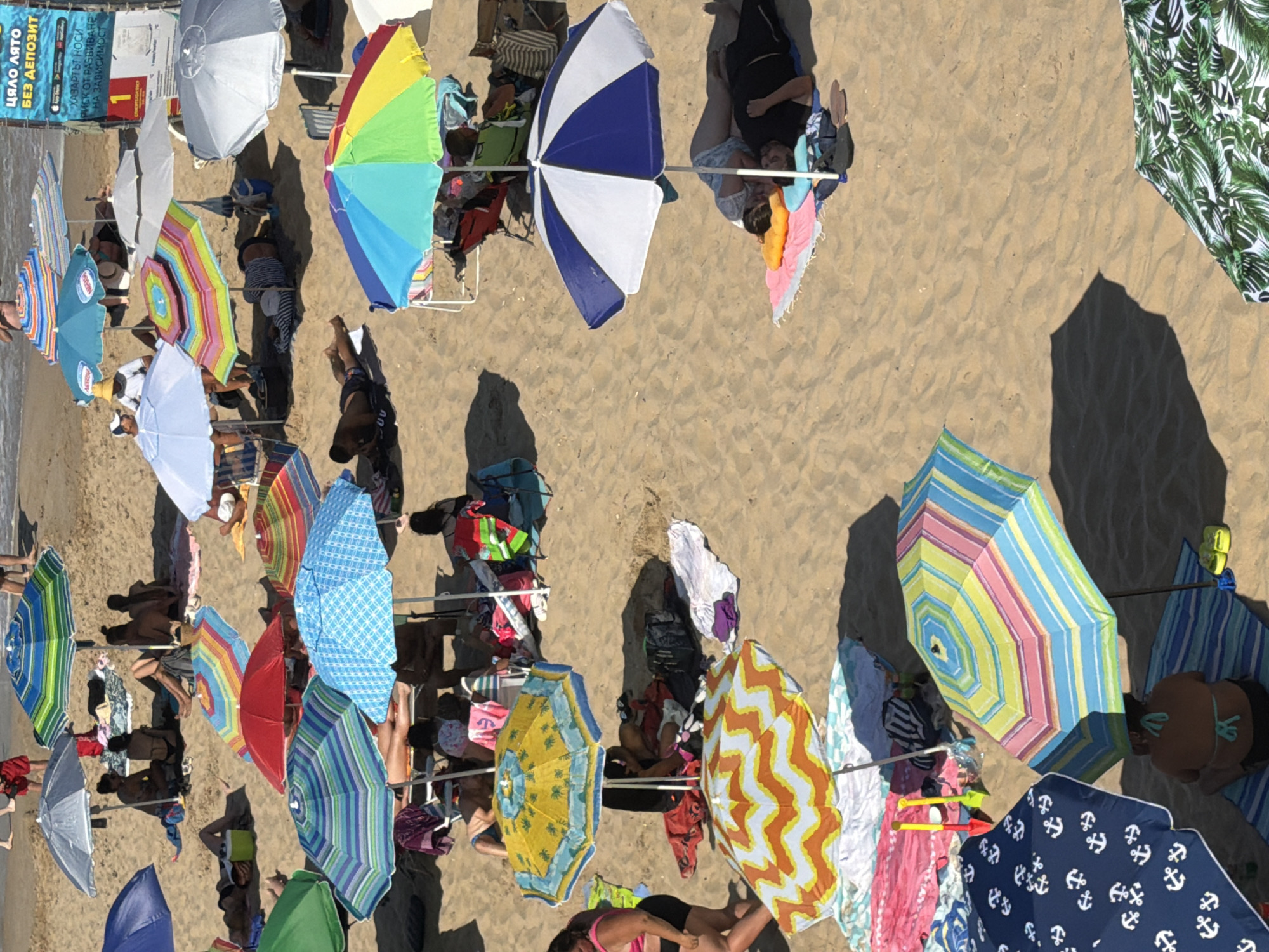 This is a high-angle, medium shot of a crowded beach in Primorsko, Bulgaria, filled with sunbathers and beach umbrellas. The sand is a light brown, and the sea is visible in the background at the top of the frame. Numerous beach umbrellas, in various colors and patterns, are spread across the sand, providing shade for people lounging on towels and beach chairs. Some individuals are seen lying down, either sleeping or relaxing, while others are seated. The scene is bustling with activity, suggesting a popular beach on a sunny day.