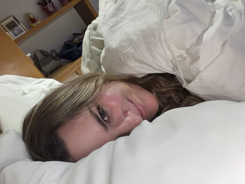 The scene is an indoor environment located in the city of San Bernardino Tlaxcalancingo, Mexico. A single female individual is depicted lying on her right side, partially obscured by bedding. Her head rests on a large white pillow that covers the left side of her face. Her right eye, exhibiting a blue iris, is visible and directed towards the viewer. Her face, characterized by light brown hair, displays a subtle smile, and her cheeks appear flushed. She is covered from the neck down by a white, wrinkled blanket or comforter. In the upper right background, a wooden furniture piece is visible, upon which a framed picture, a dark bag, and other indistinct objects are placed. The overall environment suggests a bedroom setting with ambient lighting.
