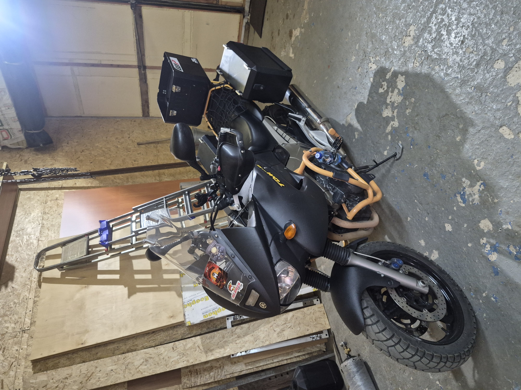 A motorcycle is parked in a garage. The motorcycle is black with an orange frame and has luggage mounted on the rear. The garage has walls made of plywood, and the floor is concrete. A ladder is leaning against the wall in the background. The location appears to be a private residence. The city is Unknown, Unknown.