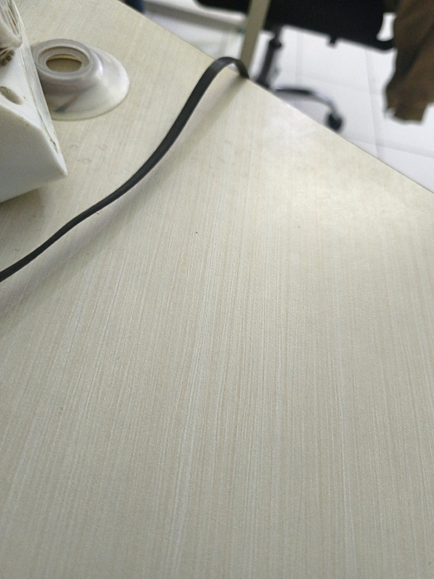 The image shows a close-up of a light-colored, textured desk surface with a black cable running across it. In the upper left corner, a portion of a white object, possibly an electronic device with openings, is visible. A clear plastic ring or base is attached to this object. The desk has a wood grain pattern. The background is blurred, showing a white tiled floor and the dark shape of a chair. There is also a suggestion of a brown cloth hanging in the upper right corner. The scene appears to be indoors, likely in an office or home setting. No specific location in Surabaya, Indonesia is identifiable from the visual information.