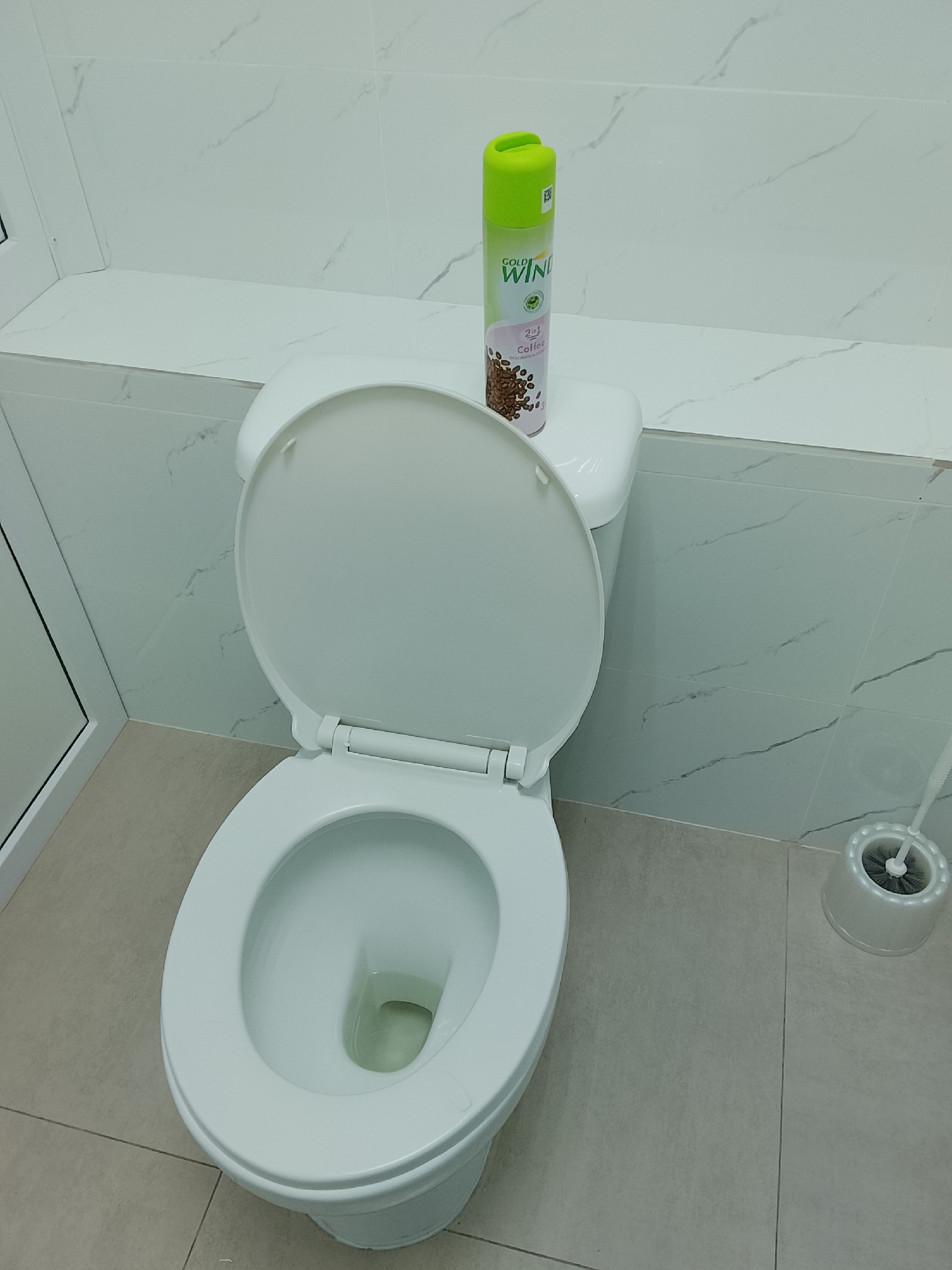 A toilet bowl with an open lid sits on a light-colored tiled floor, with a green and white spray can labeled "GOLD WIND 2 in 1 Coffee" resting on the toilet tank. To the right, a white toilet brush holder with a black brush is visible. The background consists of white tiled walls with faint grey veining. This scene is located in a bathroom in Kostanay, Kazakhstan.