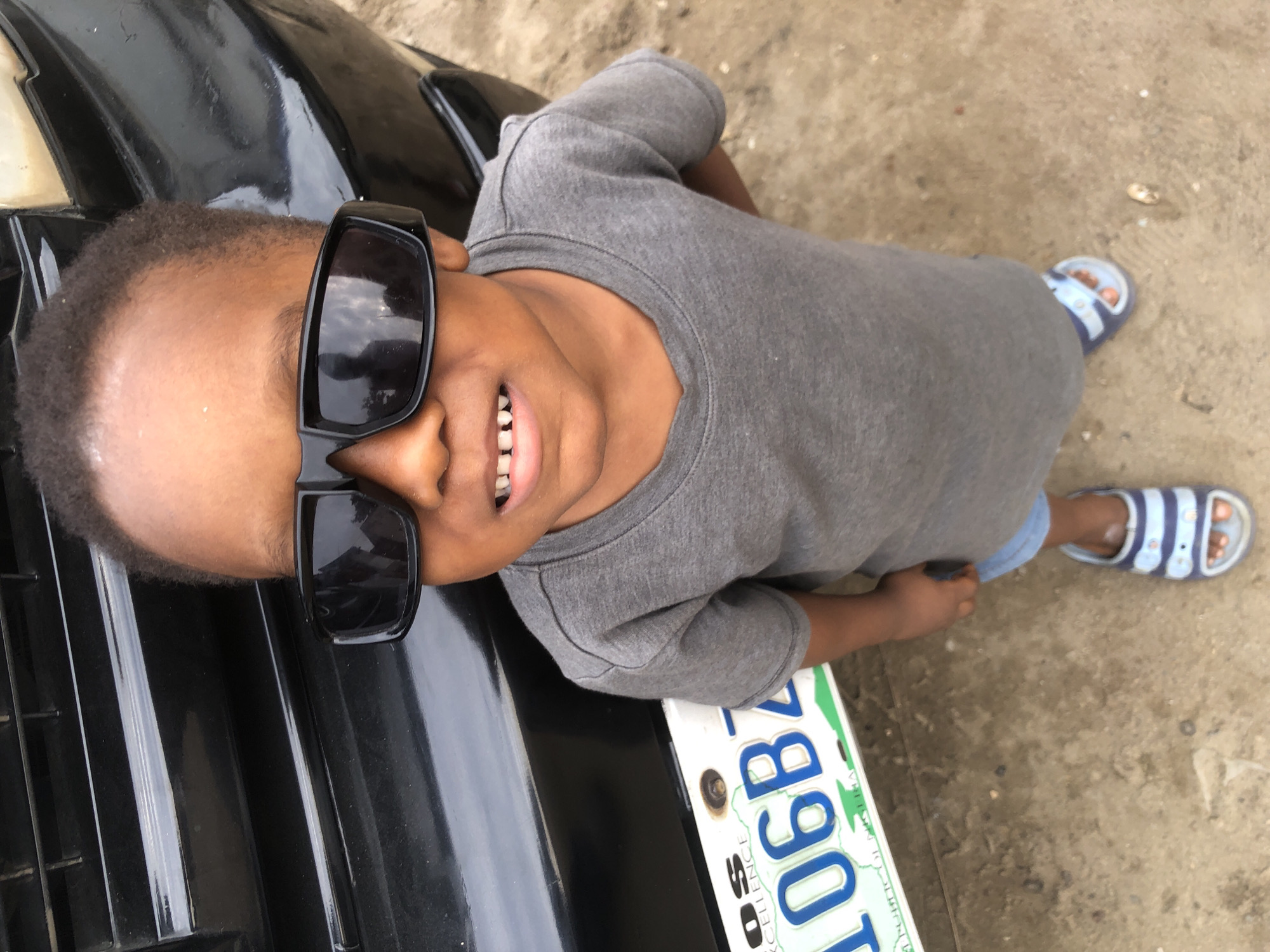 A young Black child, likely a boy given the short hair and clothing, stands facing the camera. He is wearing large, dark sunglasses and a grey t-shirt. His mouth is slightly open, revealing his teeth. He is also wearing blue and white striped sandals. The child is standing on a dirt or sandy surface, with a portion of a black vehicle visible behind him. A white license plate with blue lettering and green accents is attached to the vehicle, showing the numbers "106BZ". The overall impression is a candid outdoor photograph, possibly taken in a residential area or roadside in Ado, Nigeria.