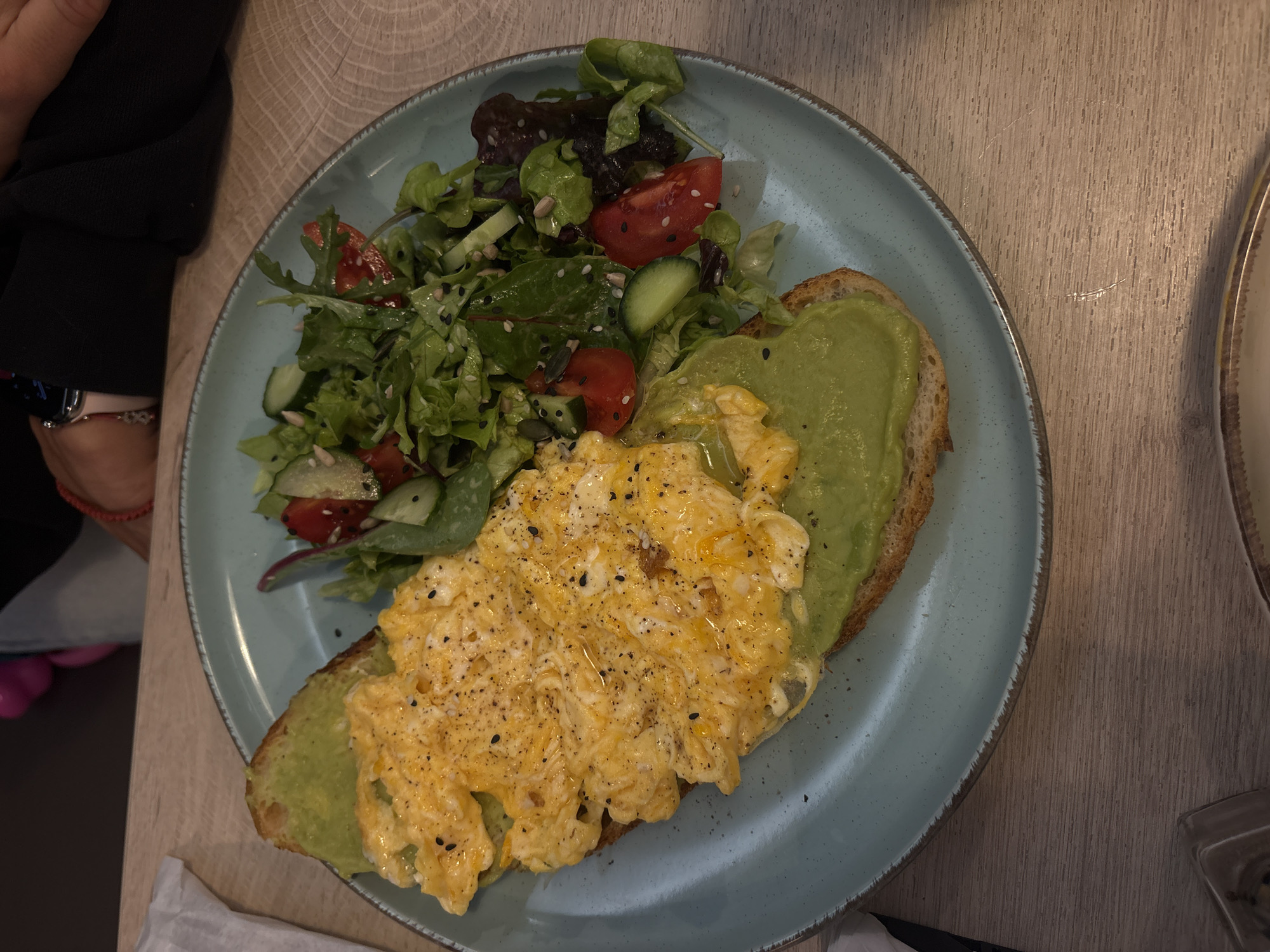 A top-down shot captures a light blue plate filled with food, set on a light wooden table. The meal consists of scrambled eggs on toast, spread with avocado, and a side salad. The salad contains lettuce, cucumbers, and tomatoes, seasoned with seeds. A person is visible on the left side of the frame, wearing a dark shirt and what appears to be a fitness tracker on their wrist. In the bottom right corner, a glass sits on the table. The location appears to be a cafe or restaurant, based on the presentation of the meal. It cannot be said for certain if it is in Varna, Bulgaria.