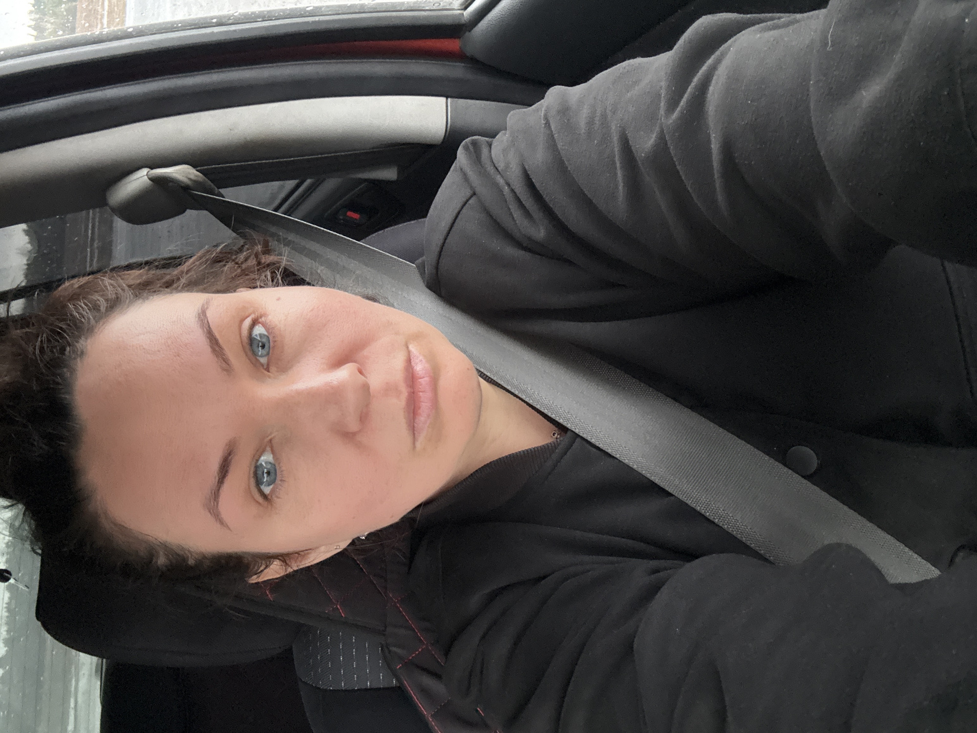 A person is sitting in a car, wearing a seatbelt and a black hooded jacket. The interior of the car is visible, including the dashboard and seats. The background shows a blurry view of the outside, possibly indicating movement or that the car is parked and it is raining. Location: Sidorovy Gory, Russia is not knowable based on image.