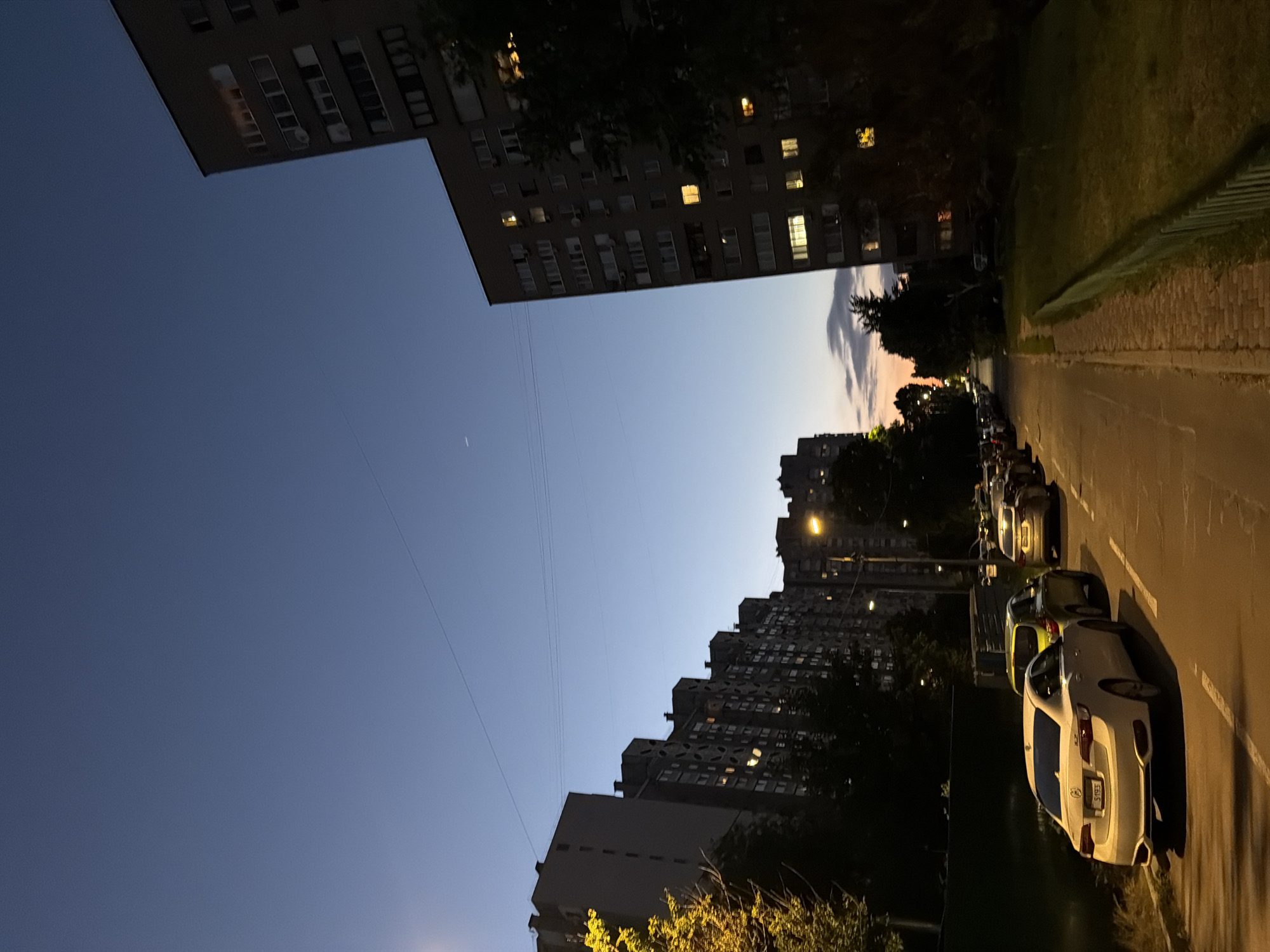 The image depicts a street scene in Kyiv, Ukraine, during dusk. Towering apartment buildings line both sides of the street, with lights visible in some windows. Several cars are parked along the side of the road. A clear, deep blue sky dominates the upper portion of the image, with a faint indication of the setting sun visible on the horizon. Trees and grass are present along the sidewalk and in front of the buildings. The overall impression is of a residential area in an urban setting as daylight fades.