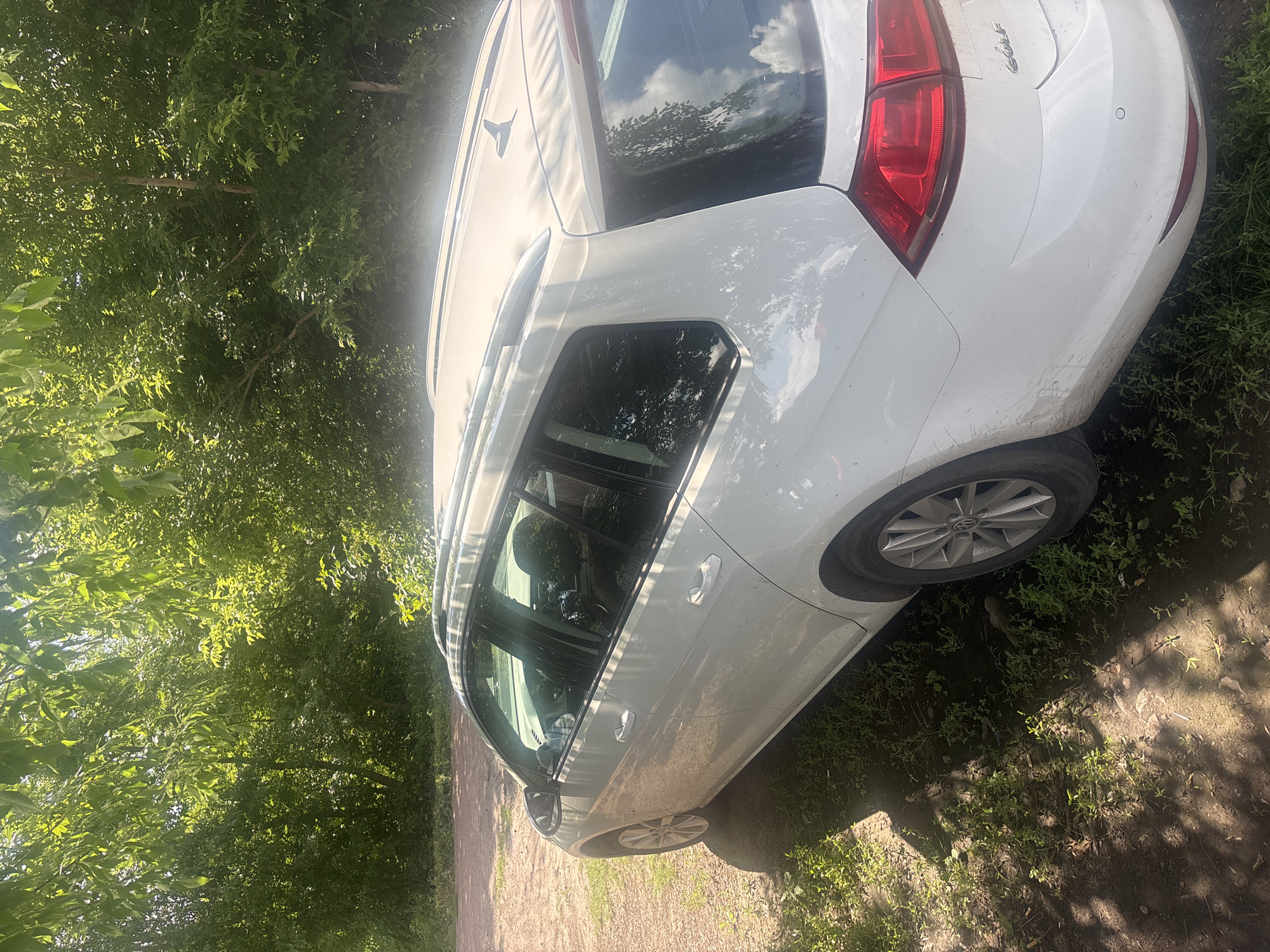 The image shows a white Volkswagen Golf station wagon parked on a dirt path surrounded by lush green trees and grass. The car is angled, with its rear and side visible. The sun is shining brightly, casting dappled shadows from the trees onto the car and the ground. The car appears to be dirty, suggesting it has been driven on unpaved roads.
The scene is set in a natural, outdoor environment, likely a rural area given the presence of trees and the unpaved path. The lighting indicates it's daytime, possibly late morning or early afternoon due to the strong sunlight and shadows. The overall atmosphere is peaceful and natural.
Notable details include the "Golf" lettering on the rear of the car. The car's rear taillights and part of the bumper are clearly visible, as is a wheel with a silver alloy rim. The car's windows reflect the sky and surrounding foliage. The presence of a small, circular parking sensor on the rear bumper is also discernible.
The image appears to be taken in Pereiaslavske, Ukraine, based on the provided location context. No people are visible in the image. The activity taking place is simply the car being parked in its current location.