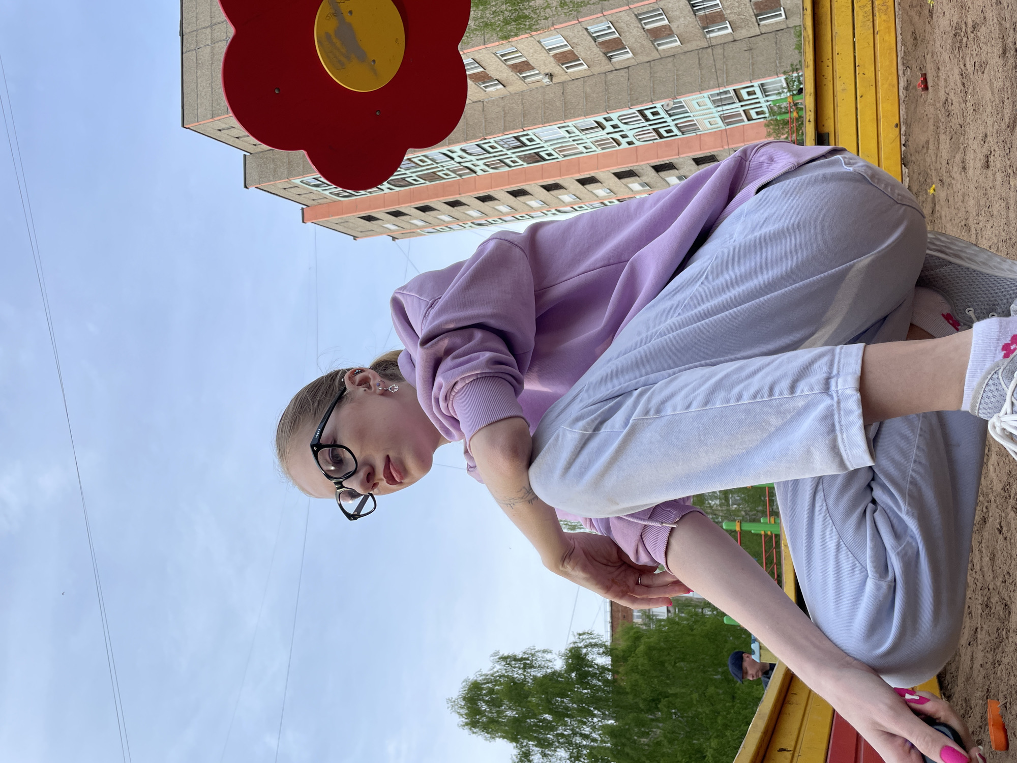 The image shows a young woman with blonde hair, wearing glasses, a light purple sweatshirt, and light-colored pants, crouching in a sandbox. She appears to be playing or interacting with something in the sand. The sandbox is part of a playground with yellow and red play structures. Behind the woman and playground, there's a multi-story apartment building. Based on the architectural style and context, the location appears to be Izhevsk, Russia. The sky is visible in the background.