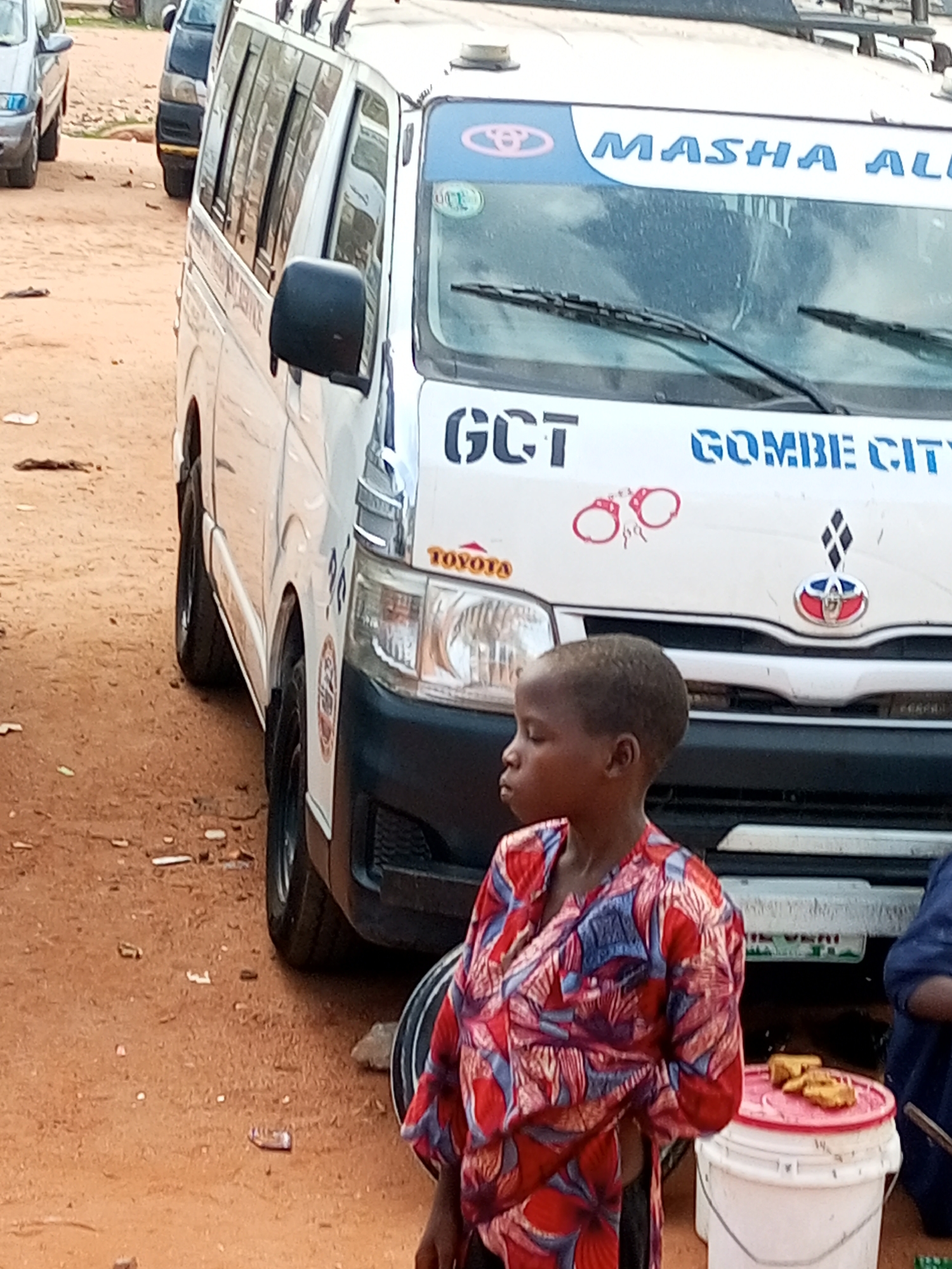 This media file captures a street scene in Gombe, Nigeria. The primary subjects are a young child in the foreground and a white commercial minibus in the middle ground. The child, with dark skin and short hair, is wearing a vibrantly patterned shirt with red, blue, and white motifs. They are looking off to the side with a neutral expression.
Behind the child stands a white minibus, a common mode of public transportation. The bus has tinted windows and appears to be parked on a dusty, dirt road. There are other vehicles partially visible in the background, suggesting a busy street or market area. A white bucket with some food items, possibly fried snacks, is placed on the ground near the child.
Visible text on the bus includes "MASHA ALL" and "GOMBE CITY" in blue lettering. The Toyota logo is also present. Red handcuff graphics are visible on the side of the bus, alongside other decals. The scene appears to be set during daylight, with natural lighting. The overall impression is a snapshot of everyday life in a Nigerian city, with elements of commerce and transportation.