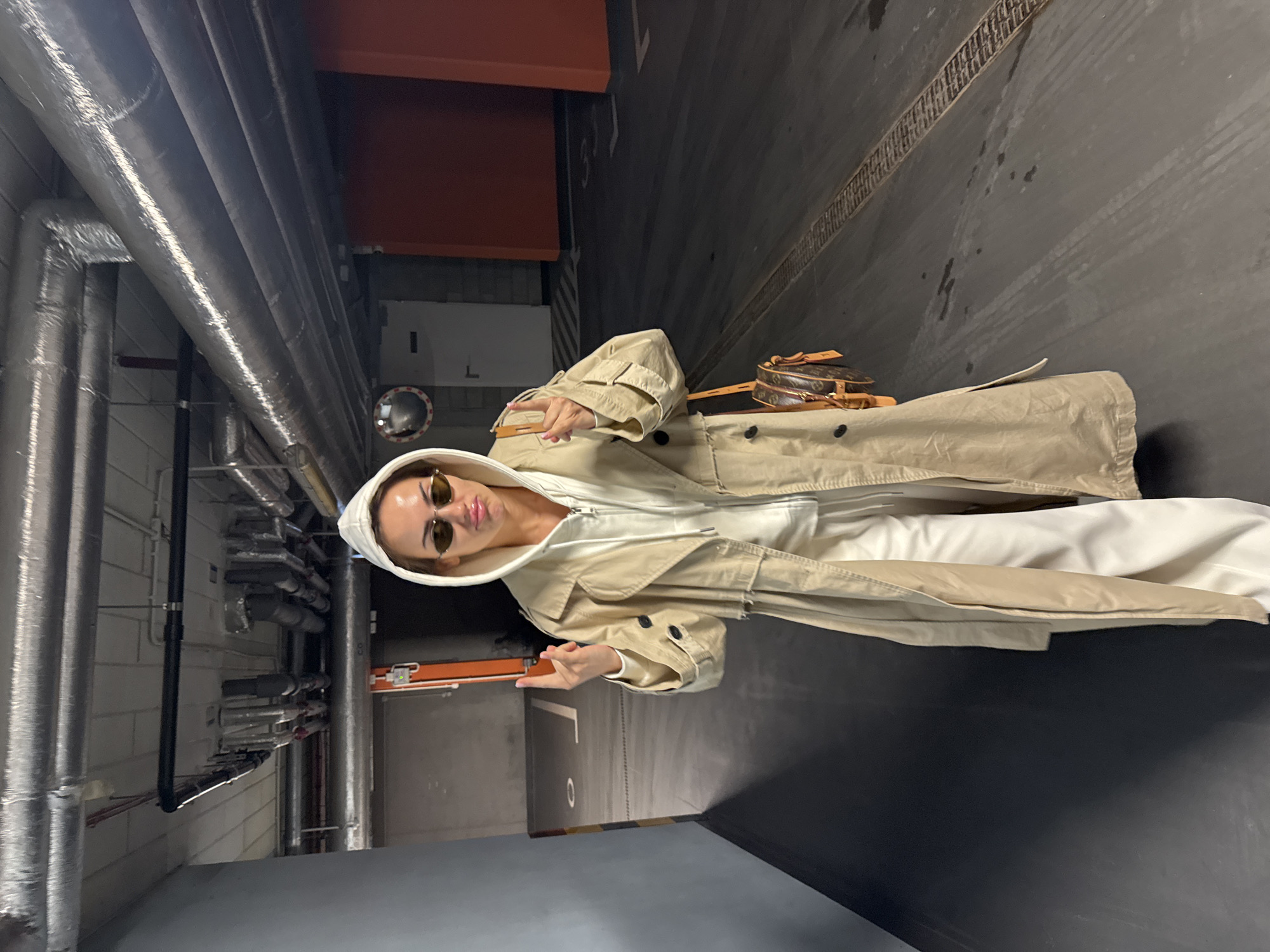 A woman stands in a dimly lit, enclosed space that appears to be an industrial or utility area. She wears a white hoodie, tan trench coat, and sunglasses, and holds her hands up in a gesture towards the camera. A brown purse rests near her waist. Behind her, exposed pipes line the wall on the left. The area on the right is a dark, smooth surface with a drain grate along the edge. The location might be a service corridor or basement in Warsaw, Poland, based on architectural styles common there.