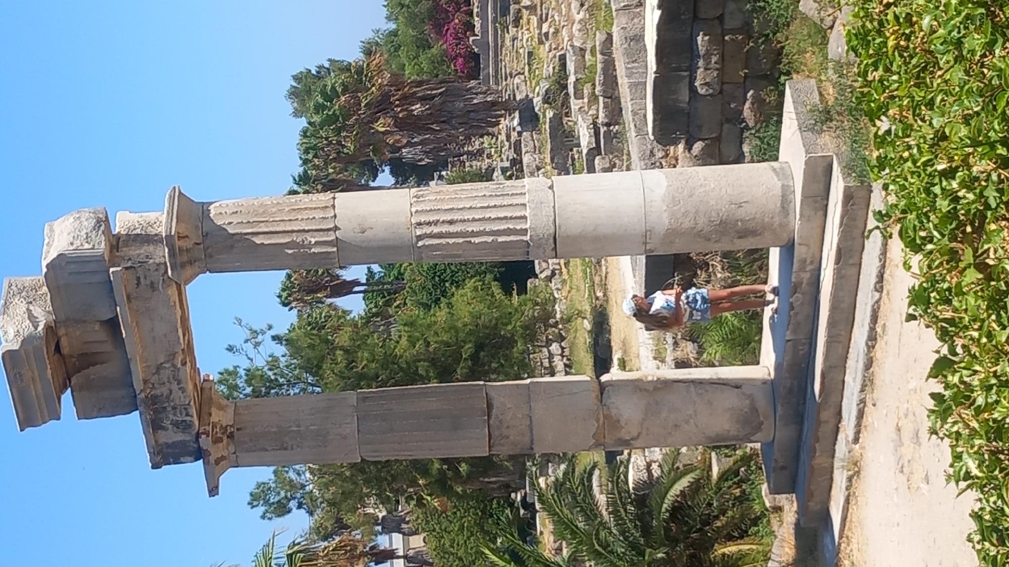 A young woman stands between two large ancient stone columns, part of an archaeological site under a clear blue sky. She is wearing a white and blue patterned top, denim shorts, and a white hat, and is looking to her right. The columns are fluted and made of weathered stone, topped with an entablature. The surrounding area is covered in ruins, with more stone structures and scattered rocks visible in the background, along with lush green trees and some pink flowers. The ground is a mix of dirt and stone. This scene is likely from the Asklepion of Kos, an ancient healing sanctuary located on the island of Kos, Greece.