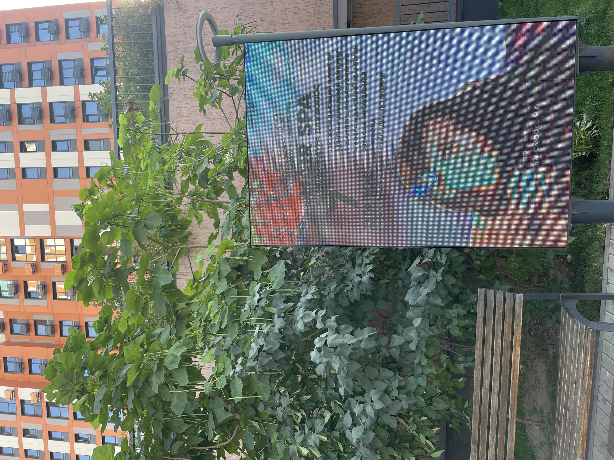 The image shows a digital billboard displaying an advertisement for "HAIR SPA" in Almaty, Kazakhstan. The billboard features a close-up of a woman's face with stylized coloring and text listing hair treatments and services. Behind the billboard, a large tree with long, dangling seed pods is prominent, and behind the tree, a modern orange and gray apartment building with numerous windows is visible. A wooden park bench is situated in the foreground to the left. The scene appears to be outdoors, likely in a residential or public area, with greenery and paving stones present.