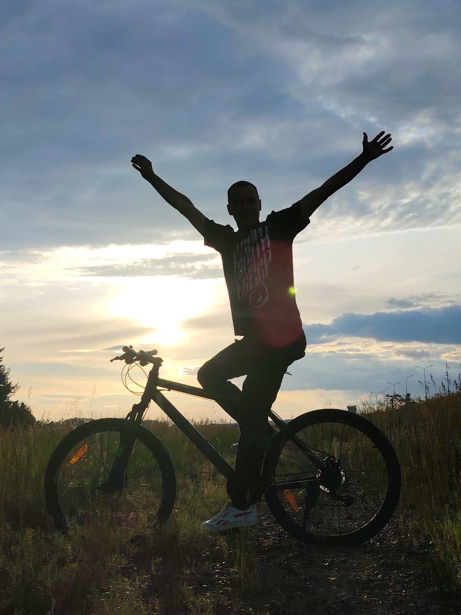 A silhouette of a person is seen on a bicycle in an outdoor setting, with the sun setting in the background. The person is standing on the pedals with their arms raised in a triumphant gesture. The sky is cloudy, with the sun peeking through, casting a warm glow on the scene. The bicycle is a mountain bike, and the person is wearing a t-shirt and pants. The setting appears to be a field or a park, with tall grass and trees in the distance. There are also streetlights visible in the background, suggesting the presence of a nearby town or city. The overall mood of the image is one of joy and freedom. The time of day is dusk, and the weather is partly cloudy. The location context is Chaikovsky, Russia. The text on the t-shirt is illegible due to the silhouette.