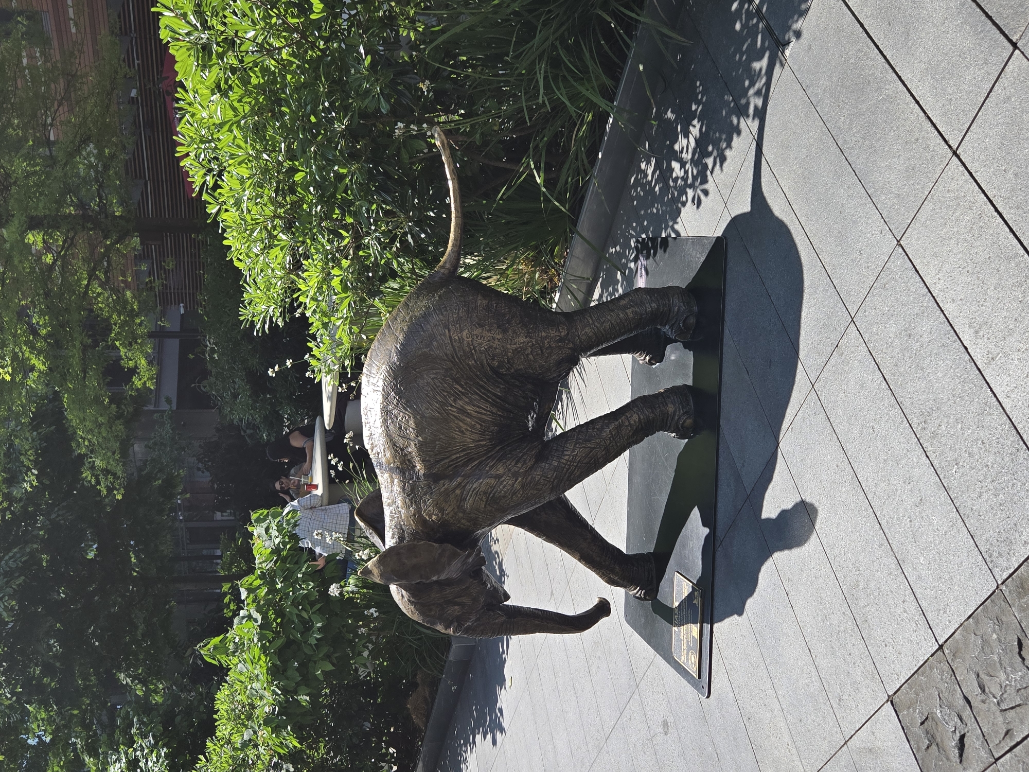 A bronze statue of a young elephant stands on a black platform on a paved walkway. The elephant has its trunk pointed down and is in motion. The walkway is made of light grey rectangular stones. To the left of the elephant is lush green foliage. In the background, blurred, are people seated at an outdoor table. A shadow of the elephant stretches across the walkway. The location is London, United Kingdom.