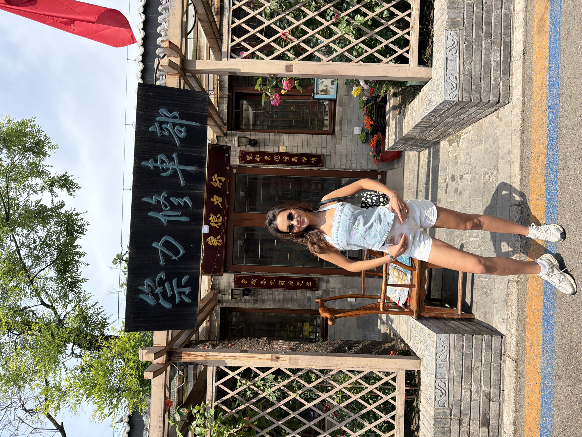 A vertical, eye-level shot reveals a woman posing in front of a traditional-style building in Dongcheng District, China. The woman, sporting sunglasses, a light blue top, and white shorts, is positioned slightly off-center to the right, smiling and holding her midsection. A small, patterned purse hangs from her shoulder.
Behind her is the building, characterized by a wooden lattice facade and traditional Chinese signage in dark green. Floral arrangements in pots add color to the entrance. A traditional wooden chair sits to her left.
To the left of the building is a tree with green foliage, and a red awning is partially visible in the upper left corner. The street in front of the building has painted blue and yellow stripes along its edge.