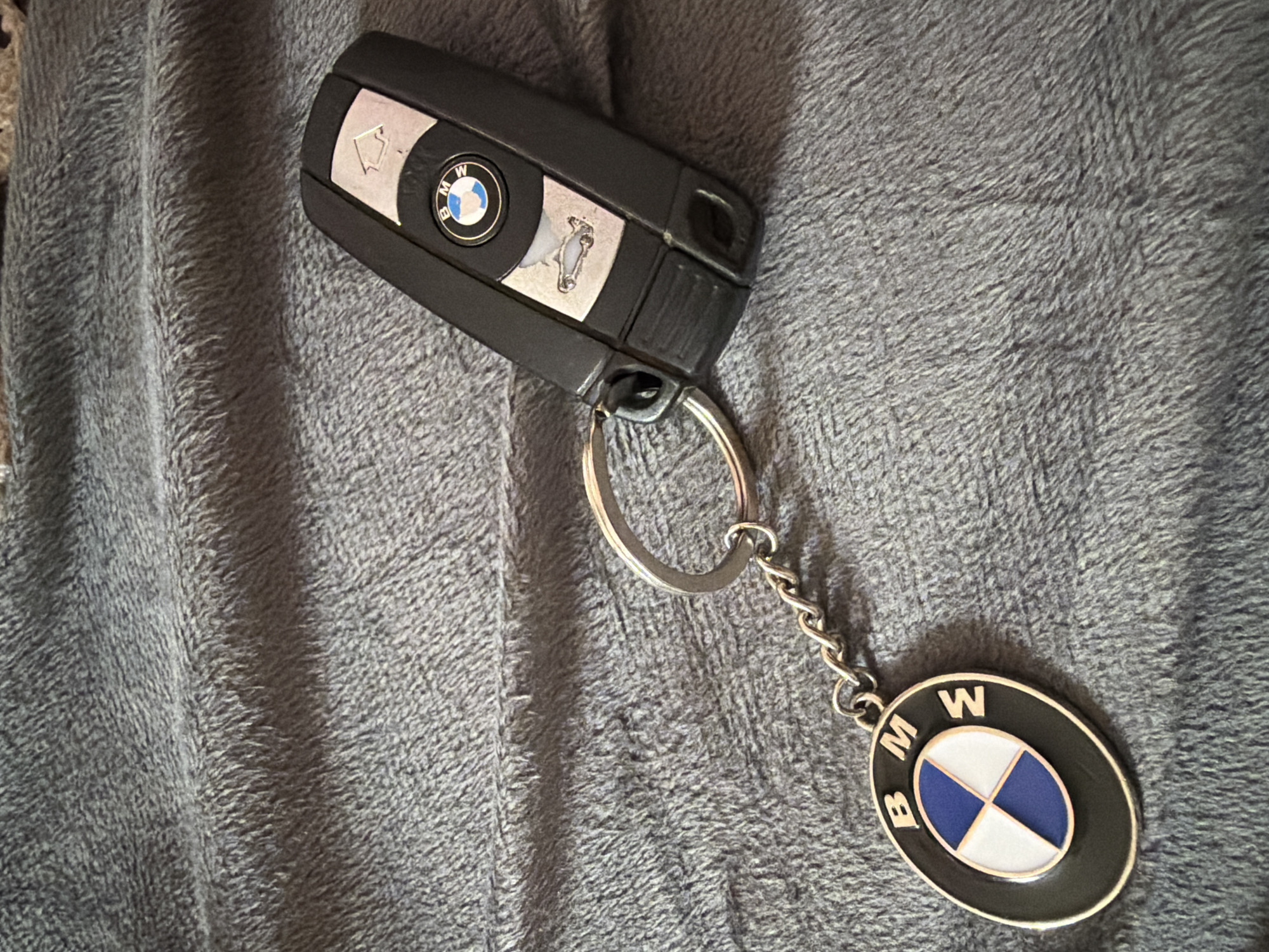 The image shows a BMW car key and keychain lying on a gray, textured surface. The key is black with silver buttons and the BMW logo on top. The keychain is silver and features an oval-shaped BMW logo pendant. The location is not specified.