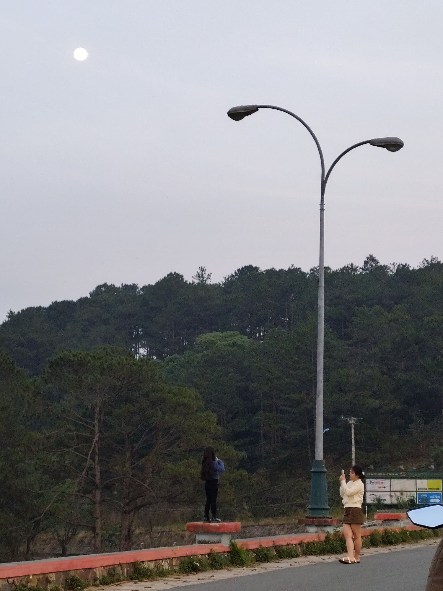 The image depicts an overcast day at an overlook point in the city of Da Lat, Vietnam. On the right of the frame, a woman in a short, brown skirt and light-colored sweater is standing on the road taking a photo. To the left of her and a bit farther away, another woman in a blue jacket and dark pants is standing on the stone wall, also taking a photo. A modern street lamp with two arms stretches above them. Behind them, a ridge covered with evergreen trees rises. The sky is grey and a bright moon is visible in the upper left corner. In the background, signs are visible behind the street lamp. The red-painted stone wall is overgrown with greenery. The road surface is asphalt. A motorcycle mirror is visible on the lower right of the image.