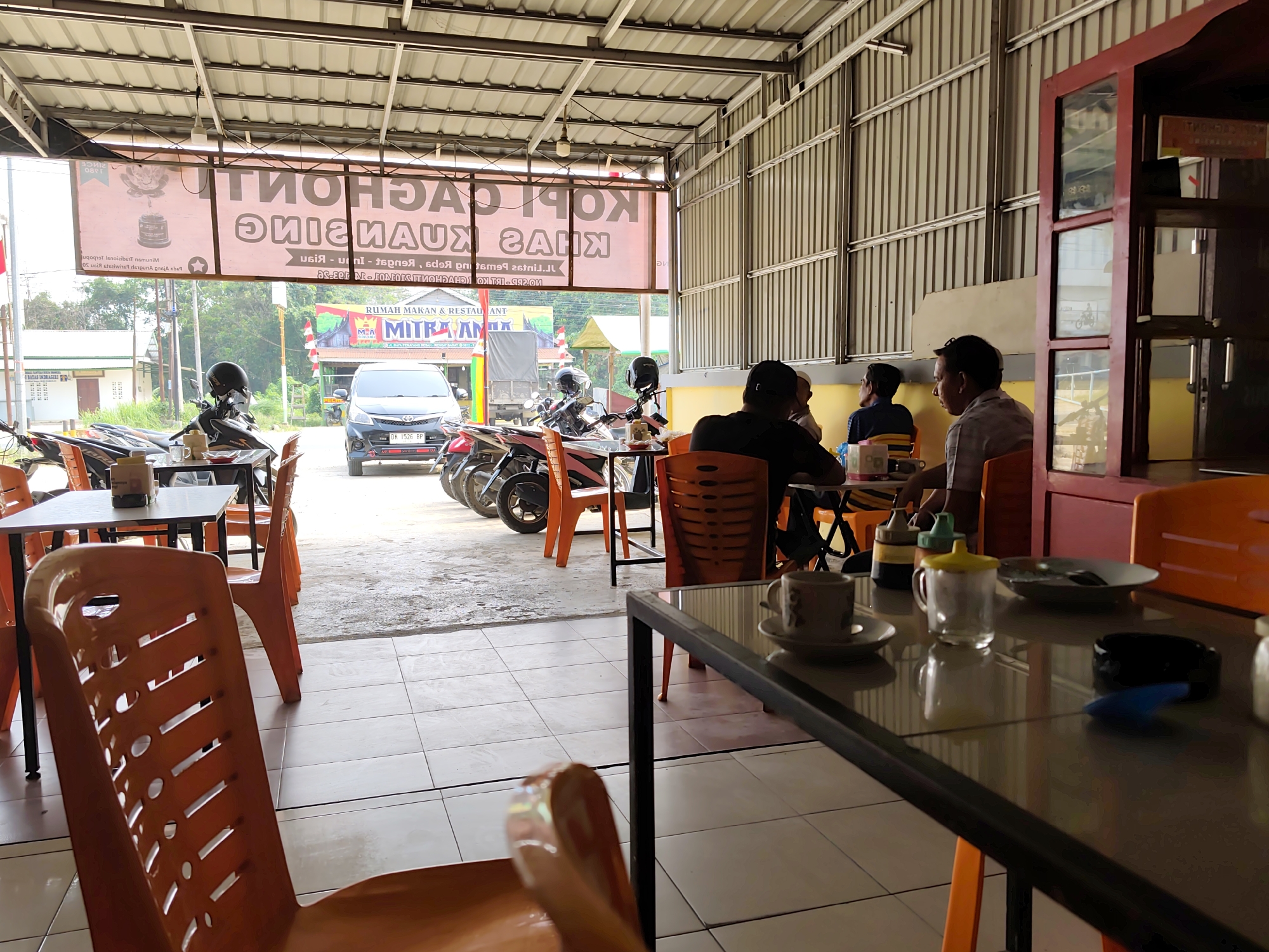 The image depicts the interior of an open-air restaurant in Pematang Reba, Indonesia. The restaurant appears to be a casual establishment, possibly a roadside eatery, with a corrugated metal roof supported by metal beams. The floor is tiled with light-colored square tiles. Orange plastic chairs are arranged around tables, some of which have glass tops.
In the foreground, a table with a glass top is visible, with a coffee cup and saucer, a glass jar, and a black ashtray on it. To the left, the back of an orange plastic chair is prominent.
In the middle ground, several people are seated at tables. Three men are the focus of attention, two of whom are engaged in conversation. One man, wearing a checkered shirt, is looking towards the viewer. Another man, in a blue shirt, is facing away. The third man, in a black t-shirt, is also facing away.
Behind the diners, several motorcycles are parked. A dark-colored car is also visible outside the establishment. A large banner with text in Indonesian, primarily pink and white, is hung across the front of the restaurant. A smaller sign with "MITRA AM" and "RUMAH MAKAN & RESTAURANT" is also visible. The environment outside suggests a warm climate, with lush green vegetation in the background. The overall impression is that of a bustling, everyday dining scene in a tropical location.