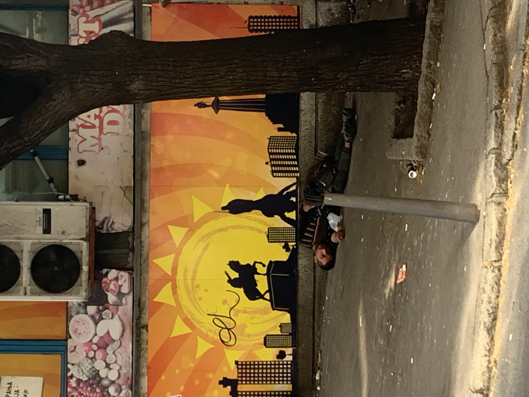 A man lies sleeping on a sidewalk next to a metal pole. He has dark hair and is wearing dark clothing and sneakers. Next to him is a backpack. Behind the man is a large advertisement featuring a stylized depiction of Belgrade, Serbia, with silhouettes of buildings and people against a yellow and orange sun. A large tree trunk and branches obscure a portion of the advertisement and adjacent wall, where an air conditioning unit and fruit advertisement are visible. The location is likely in Belgrade, Serbia, given the artwork of the advertisement.