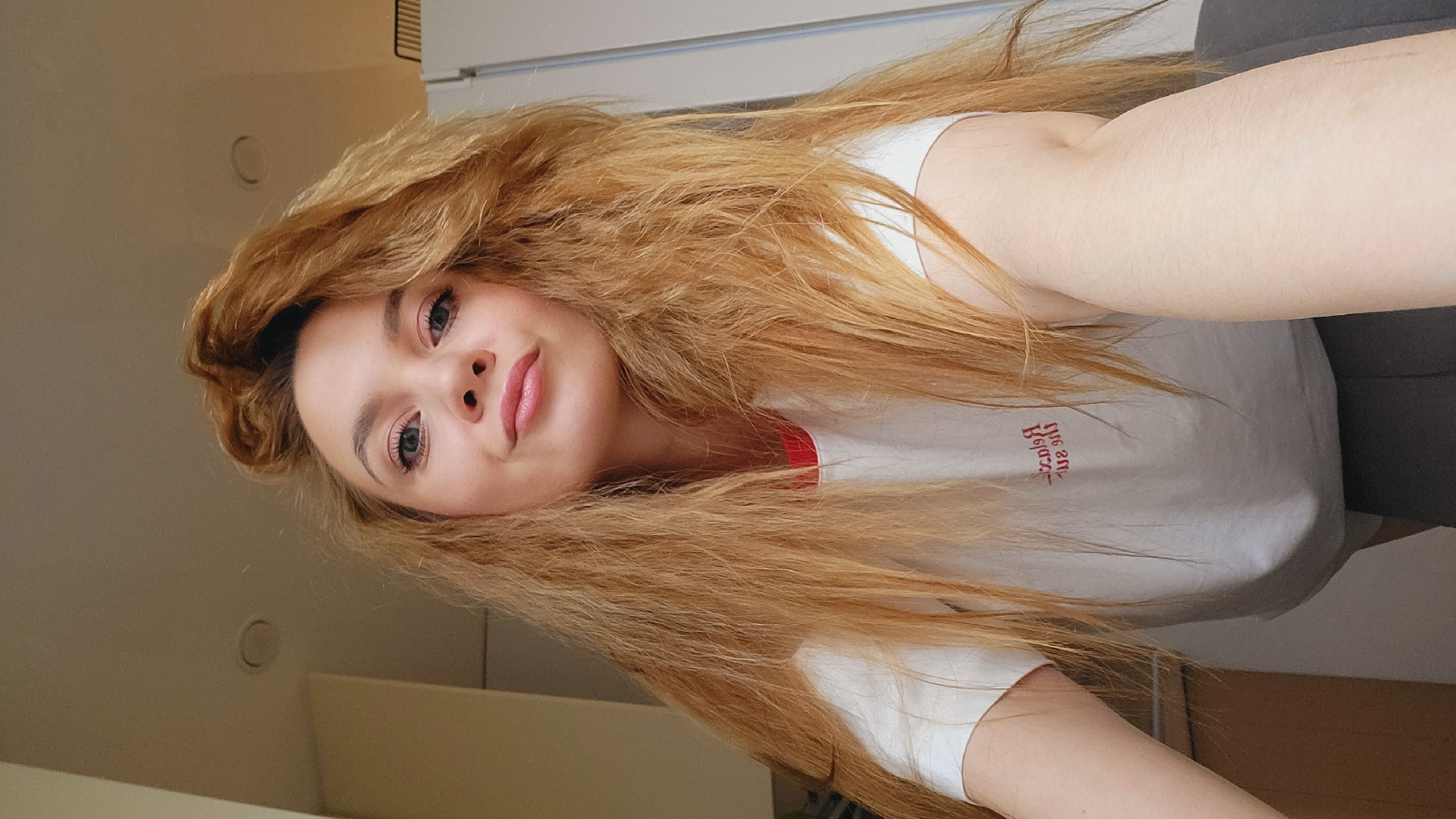 A young woman with long, wavy, light brown hair is depicted in a selfie. She has fair skin and is wearing a white t-shirt with a red trim. The location is indoors. The city of Chaikovsky, Russia, cannot be determined from the image.