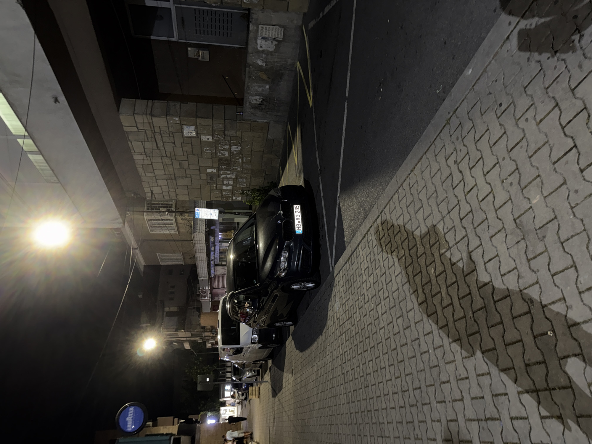 At night, on a city street in Yambol, Bulgaria, cars are parked along the side of the road. A dark sedan with a license plate reading "HB AO 285" is in the foreground, positioned parallel to the sidewalk. Behind it, a white van and several other cars are also parked. The sidewalk is paved with interlocking grey bricks. A bright street light illuminates the scene, casting long shadows across the sidewalk. The street appears to be in a commercial or residential area, with buildings visible in the background. Some people are walking on the sidewalk further down the street. A blue circular sign with the word "LAVAZZA" is visible on the left side of the image. The street is asphalt.